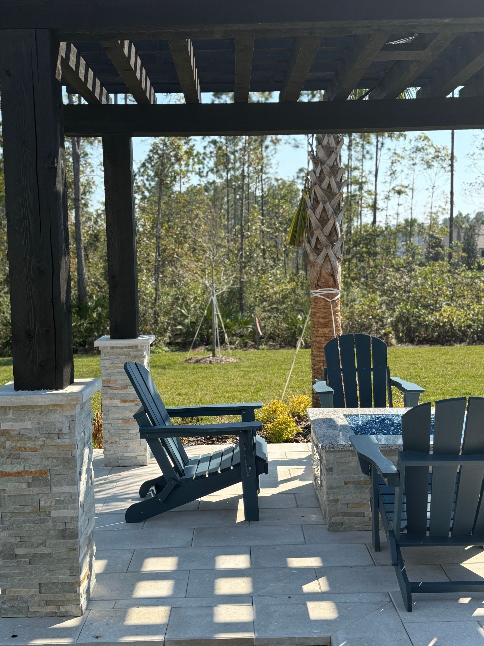 Outdoor seating area under a pergola with blue chairs, stone columns, and a grassy backyard.