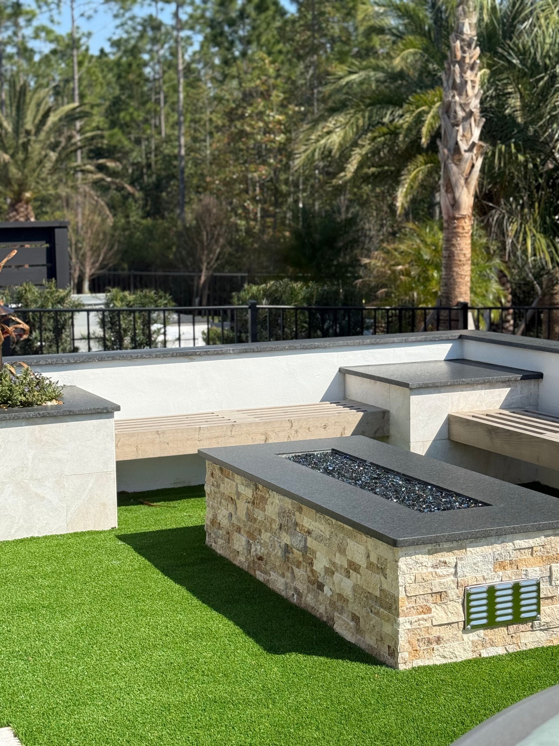 A modern outdoor patio with a stone fire pit, concrete benches, and green turf, surrounded by trees.