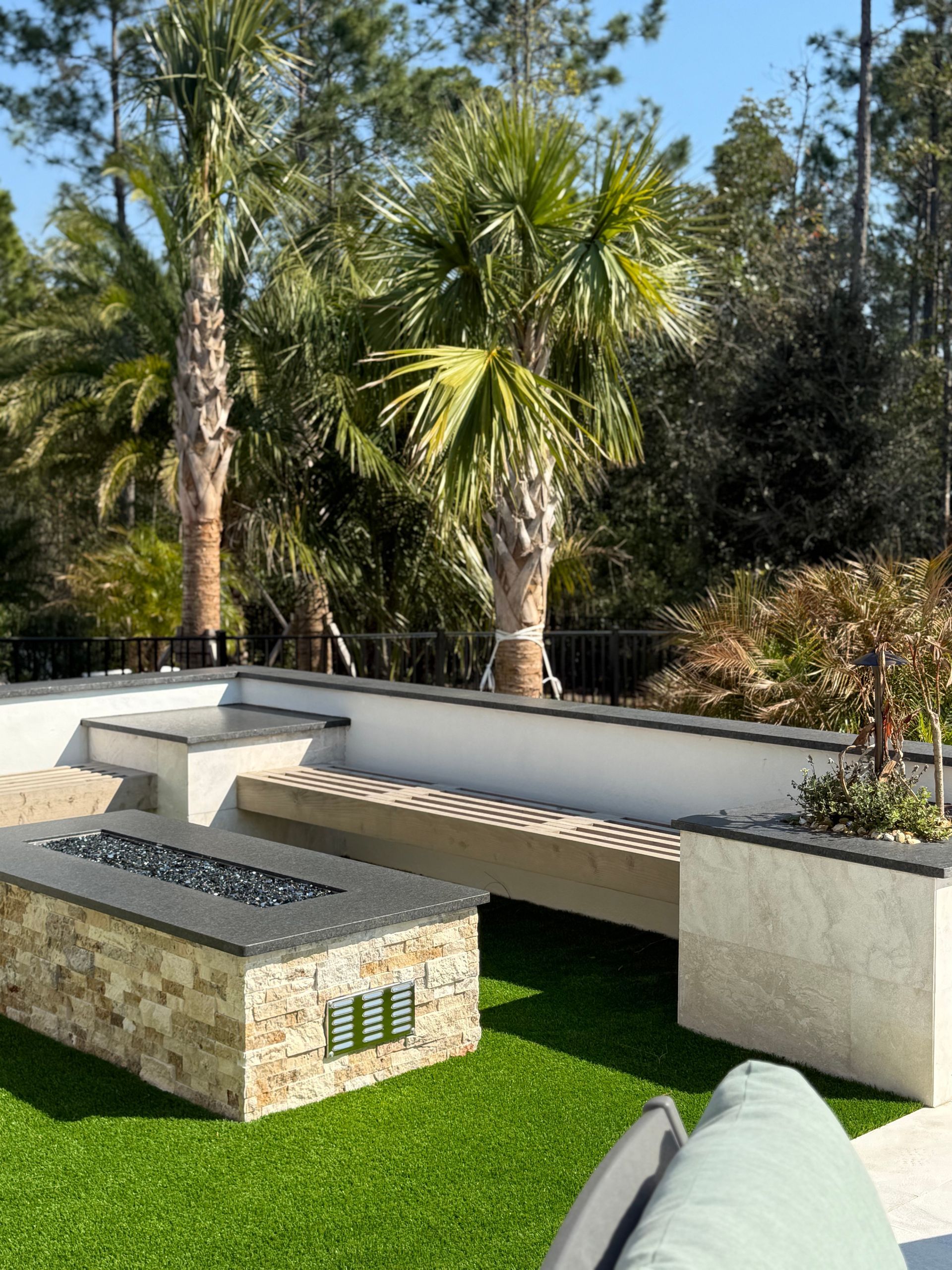 Outdoor patio with fire pit, bench, and palm trees. Green turf, white walls, and stone accents.