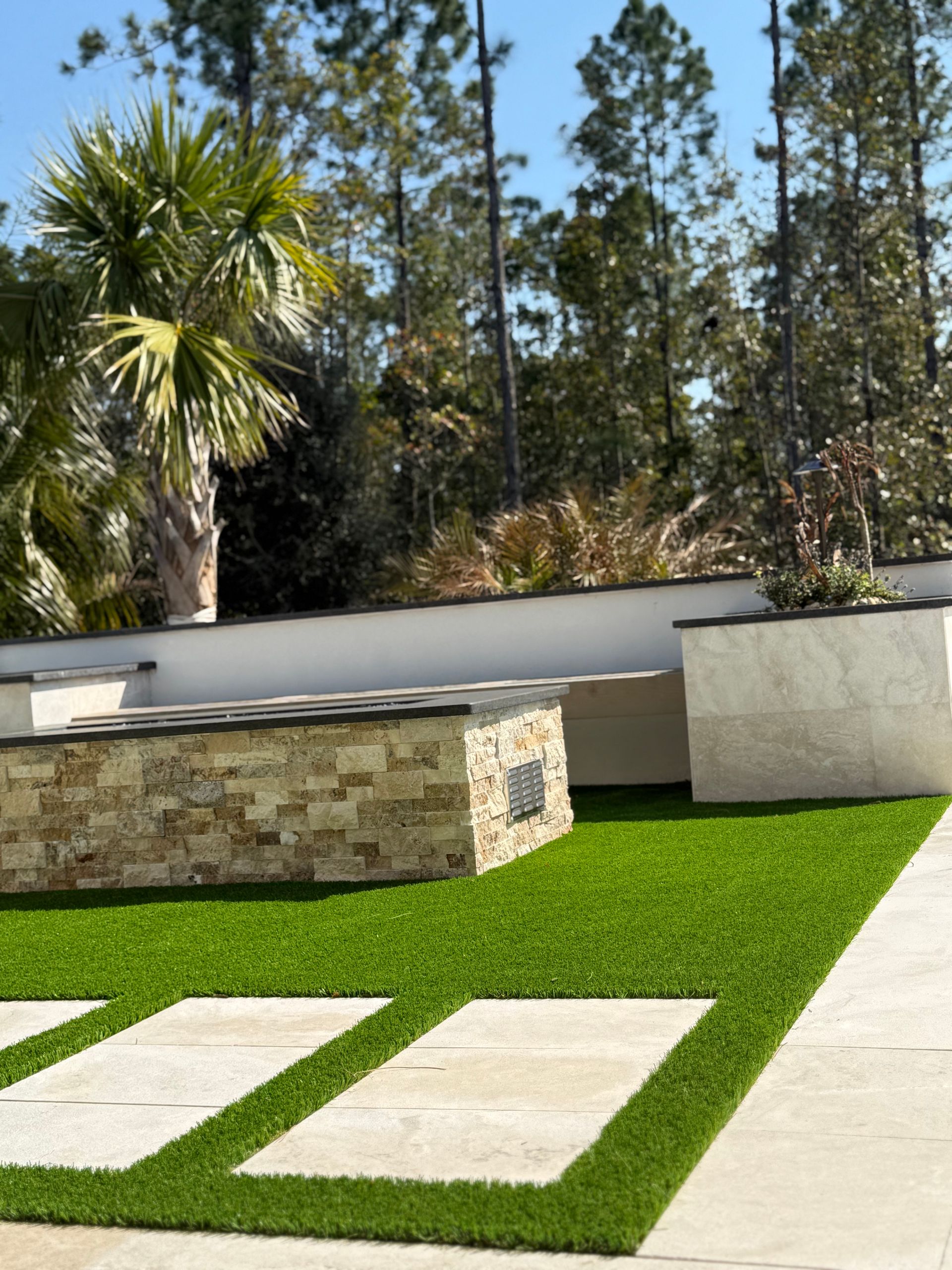 Artificial turf yard with stepping stones and a stone fire pit, greenery, and trees in the background.
