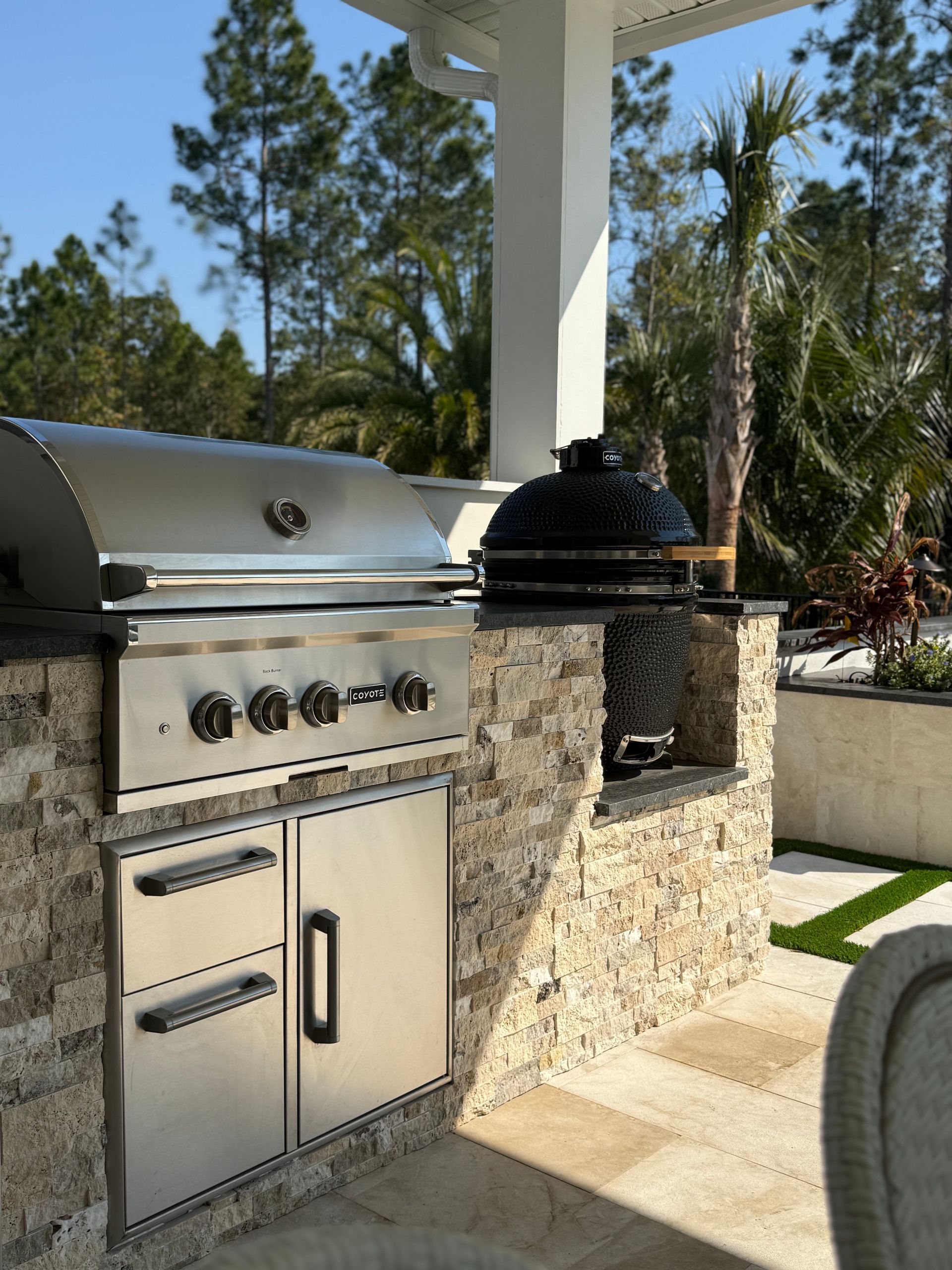 Outdoor kitchen with stainless steel grill and black ceramic smoker on stone.