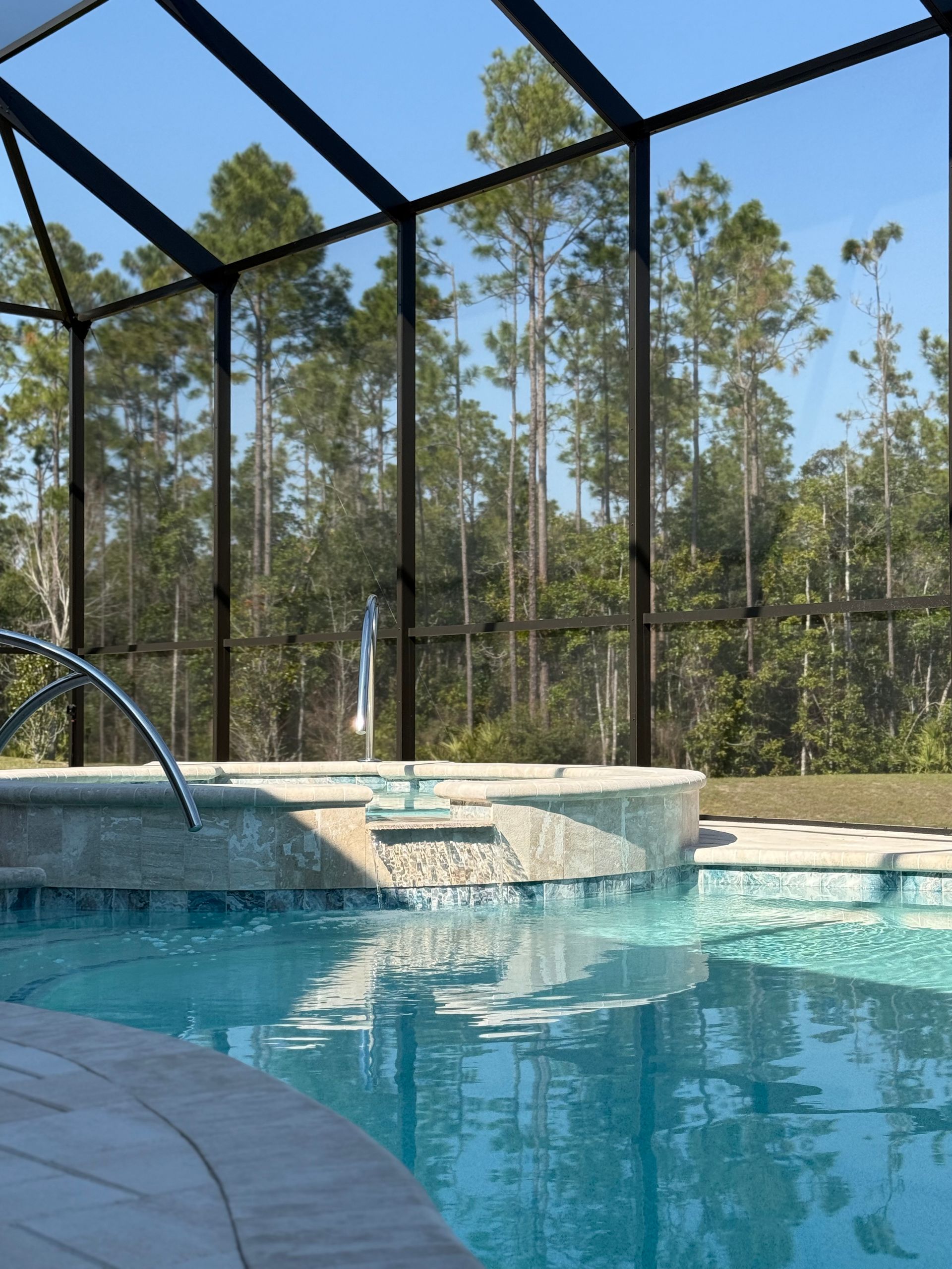 Pool with waterfall feature, enclosed by screen, surrounded by trees under blue sky.