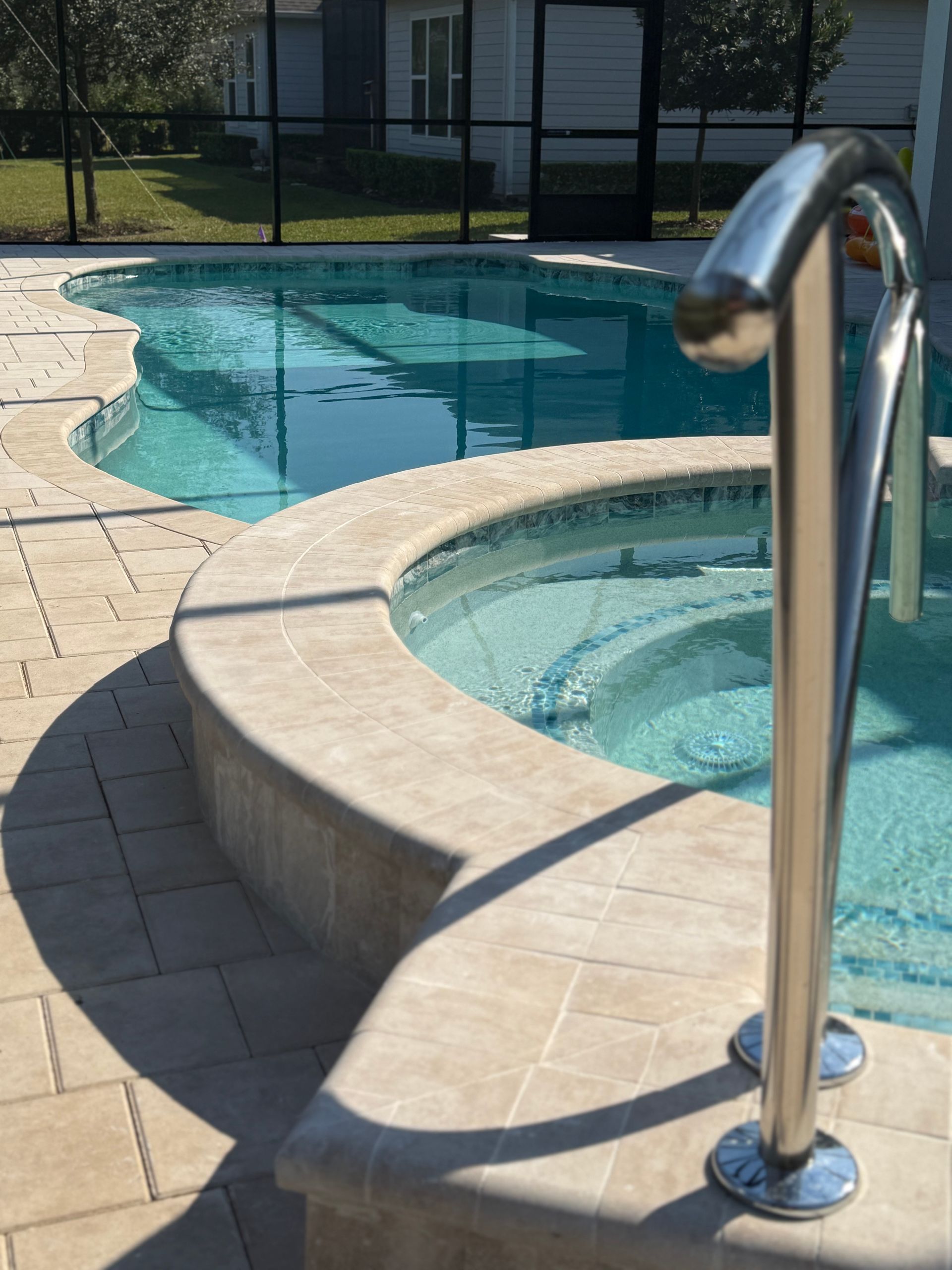 Pool with a light-colored stone surround, featuring a hot tub and metal handrail. Sunny outdoor setting.