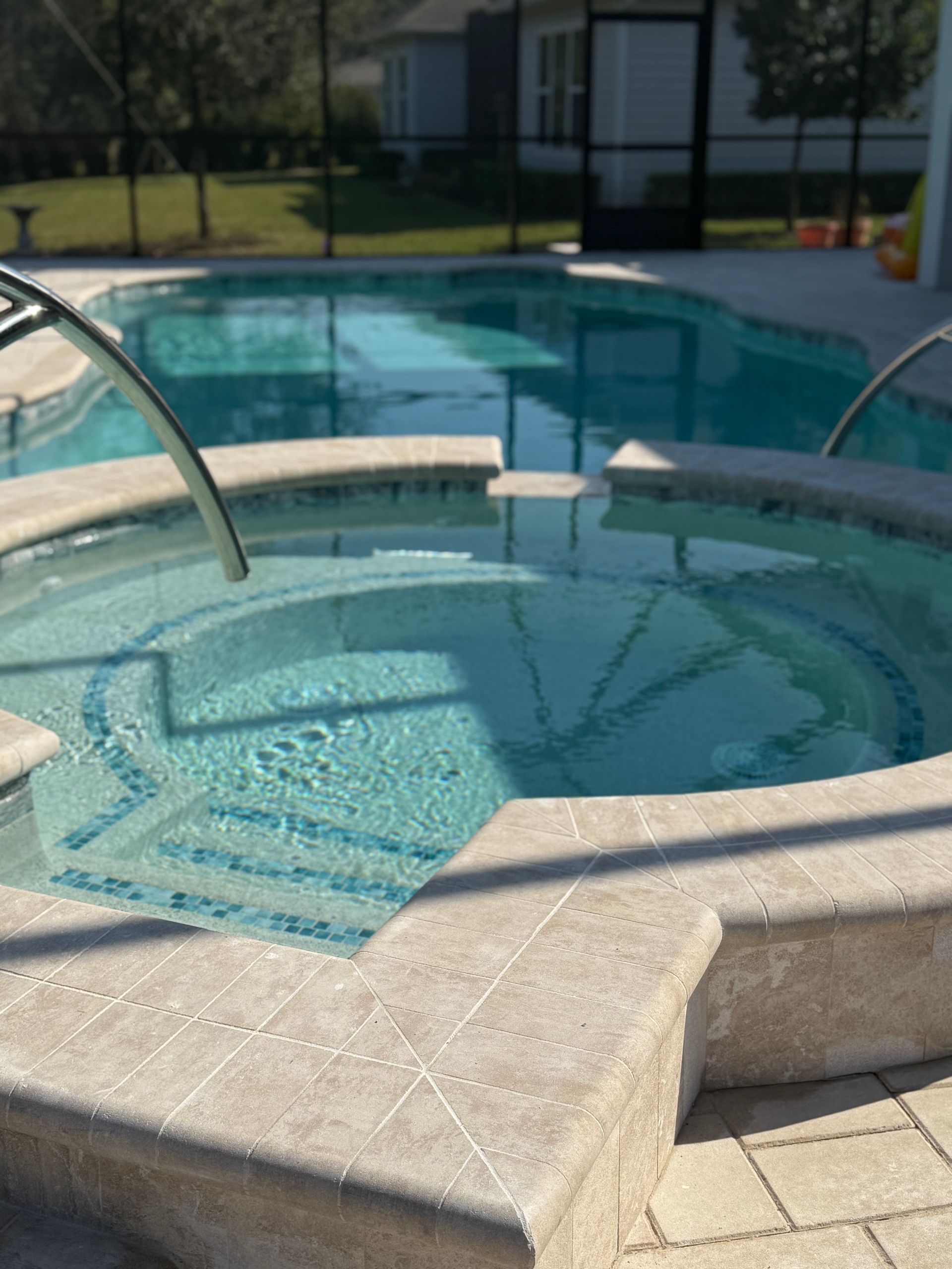 A round hot tub and pool with clear water, surrounded by beige tile, in a sunny outdoor setting.
