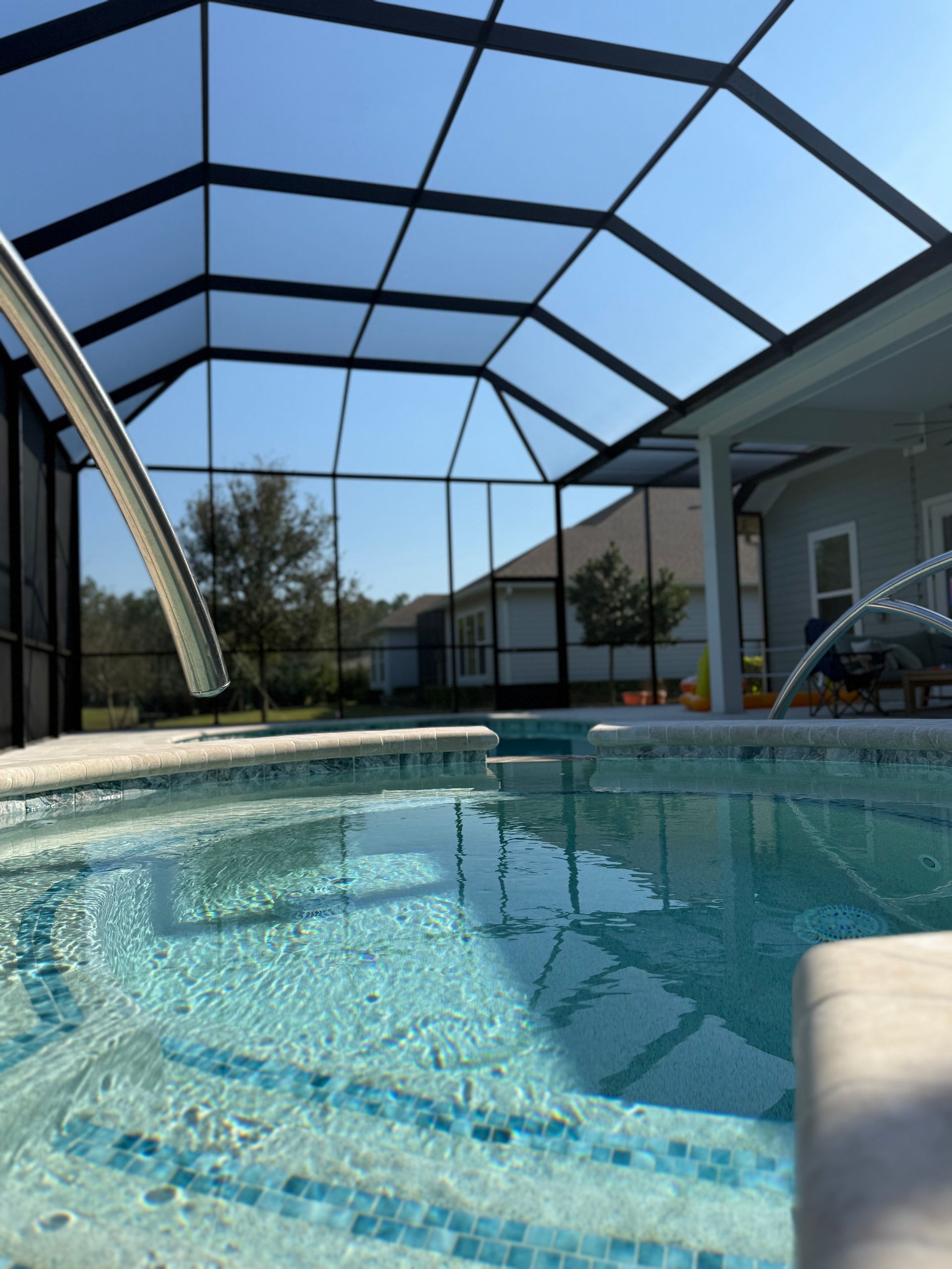 Pool under a black screened enclosure with a blue sky, surrounded by a home and trees.