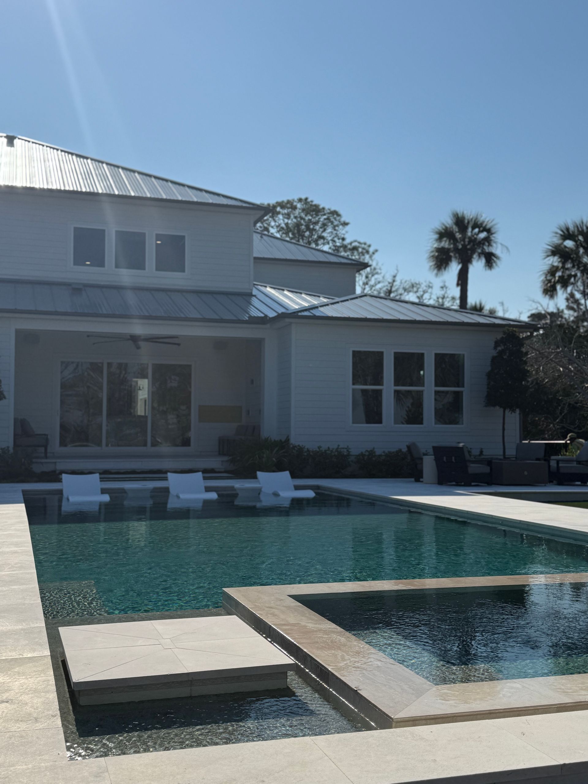 White house with pool; blue water, white lounge chairs in pool, sunny day.