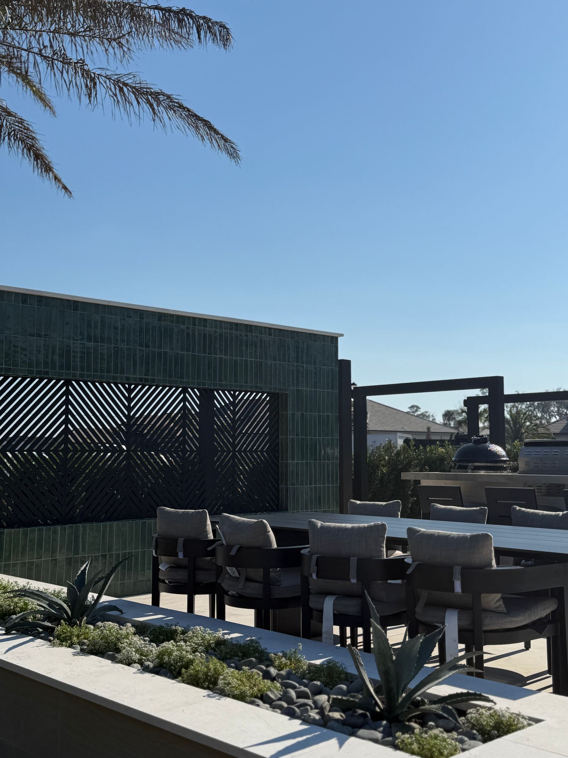 Outdoor dining area with table, chairs, and textured green wall, under a clear blue sky.