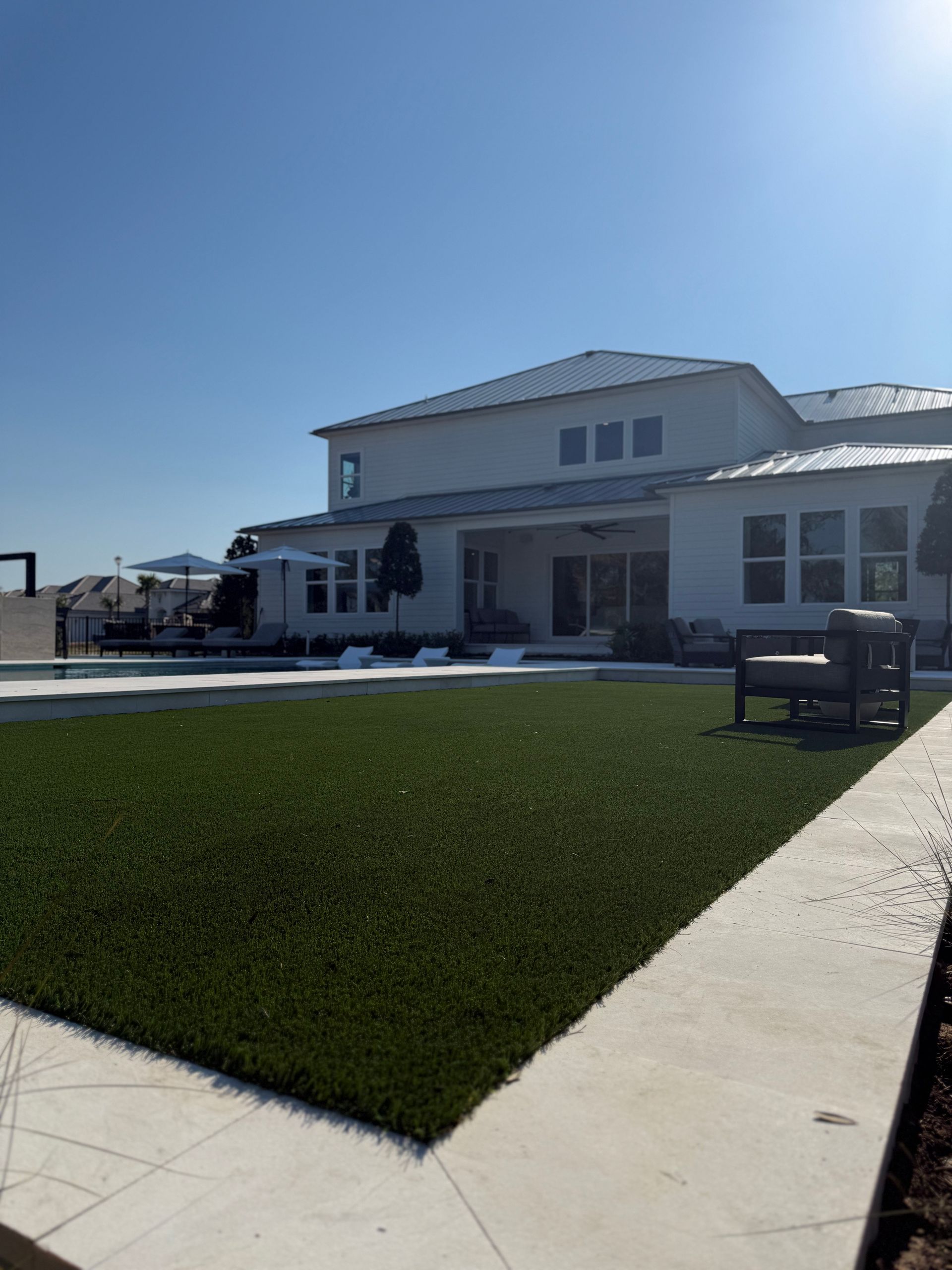 Backyard with green turf, patio furniture, swimming pool, and a two-story white house under a clear blue sky.