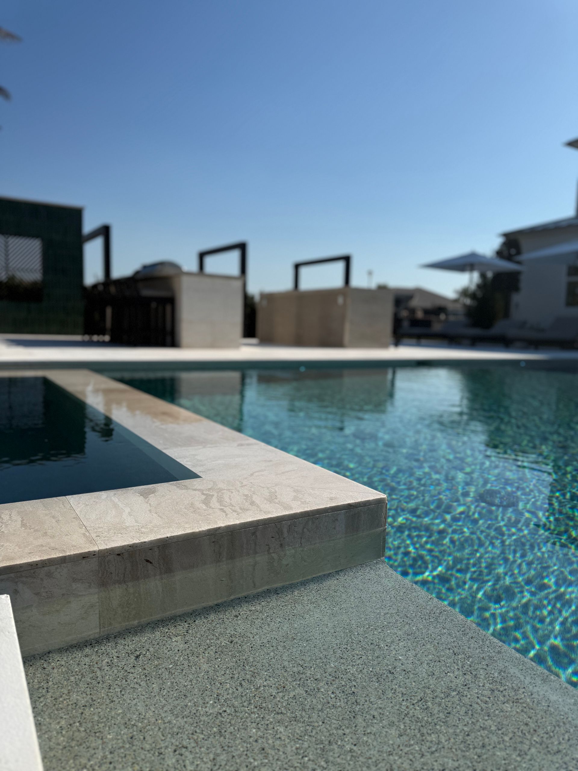 Poolside view of a rectangular pool with blue water and concrete edging, clear sky in the background.