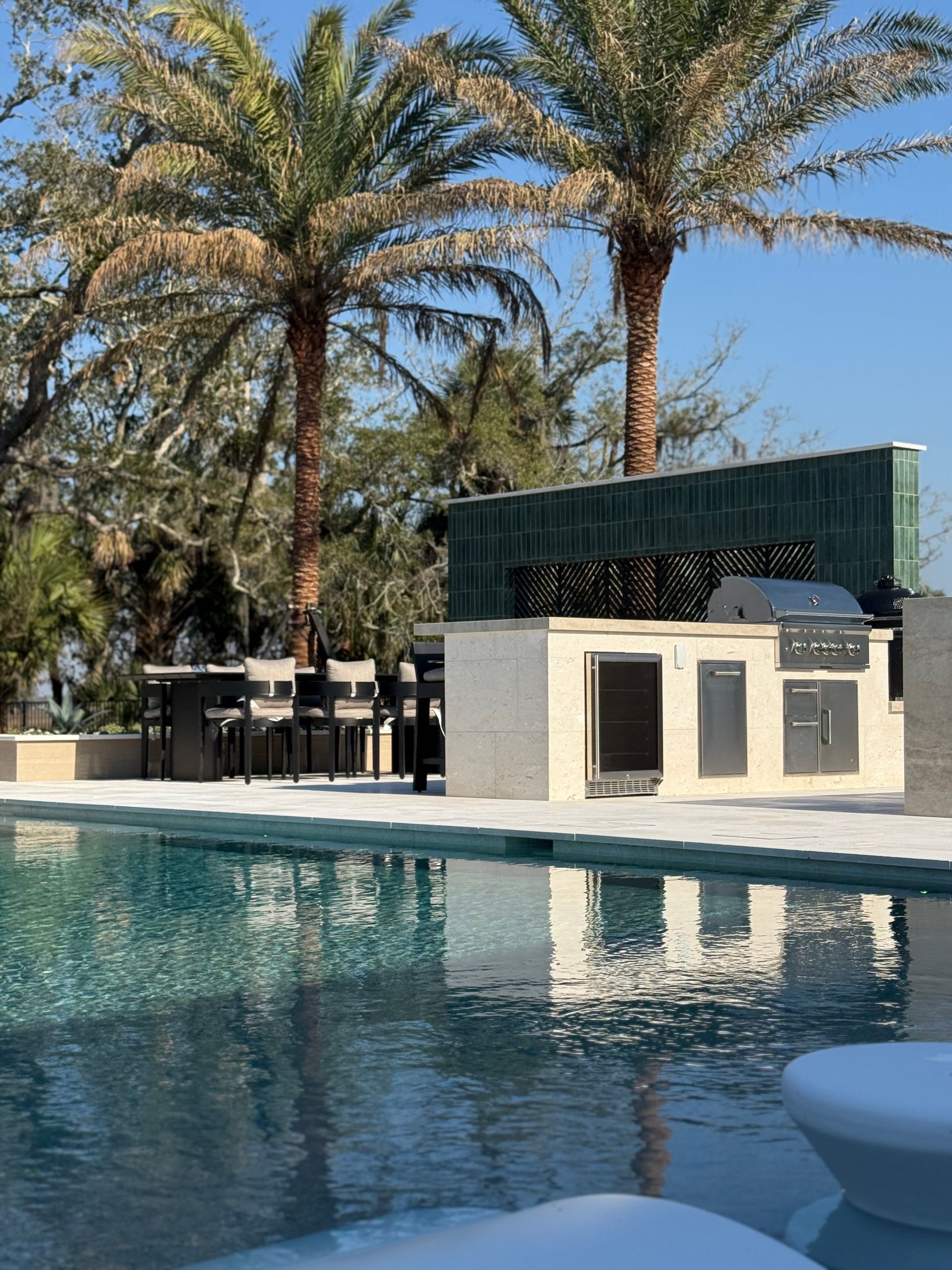 Outdoor kitchen by a pool, palm trees in the background. Sunny day.