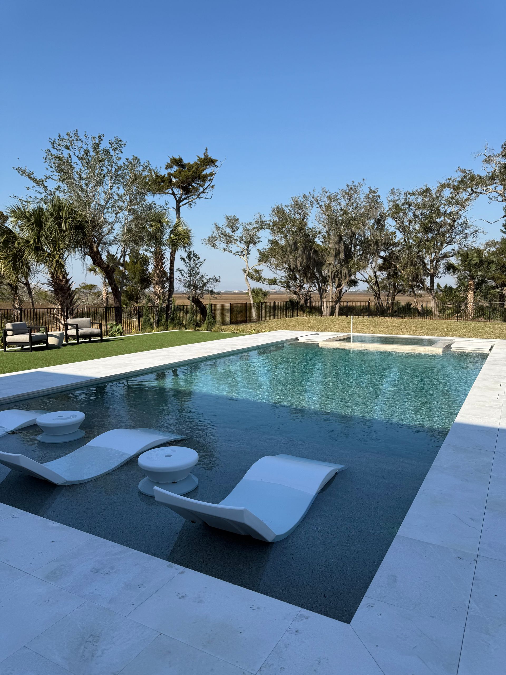 Pool with white chaise lounges, surrounded by a stone patio and green lawn, under a clear blue sky.