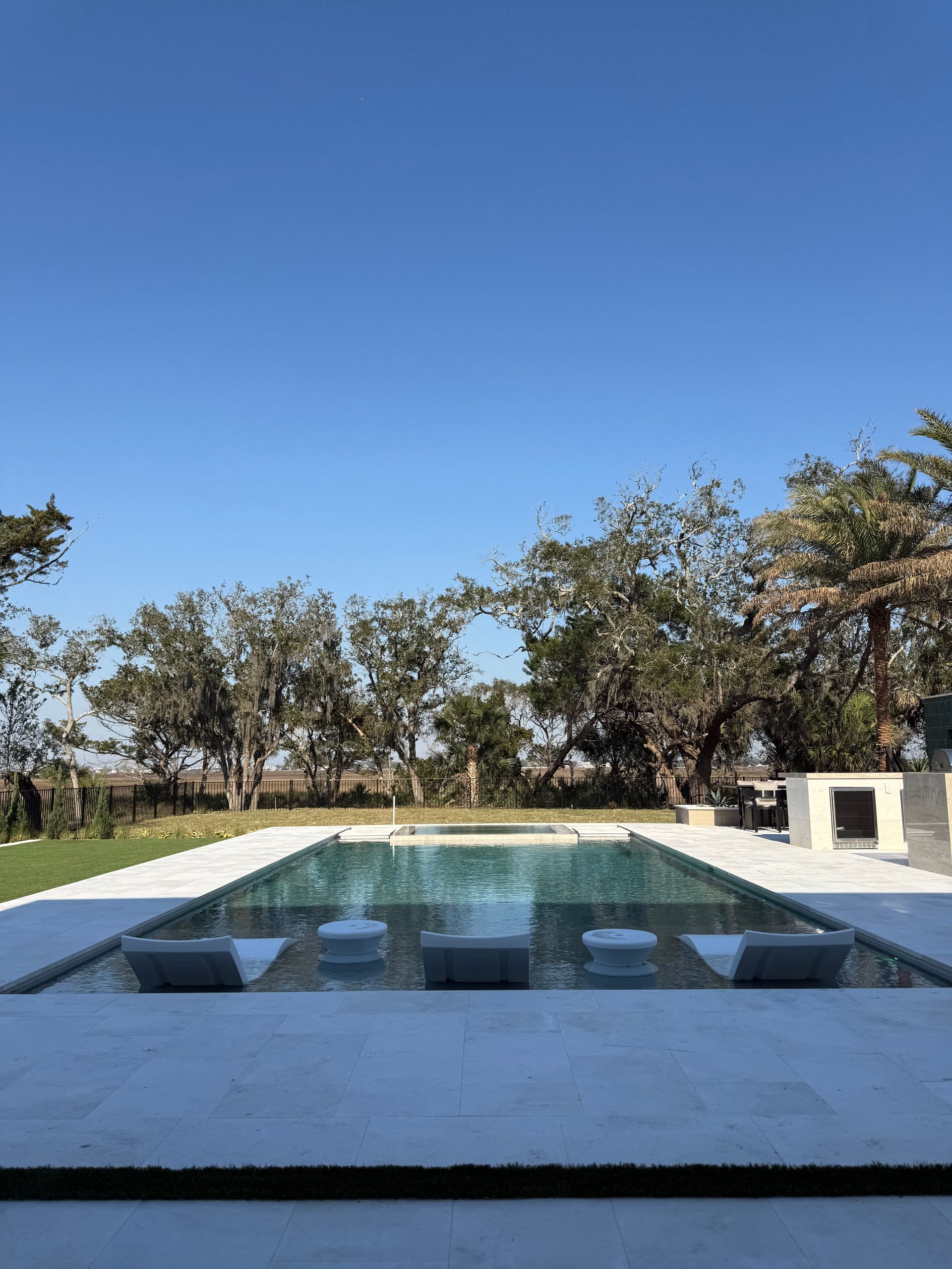 Pool with submerged seating, surrounded by stone patio, trees, and a clear blue sky.