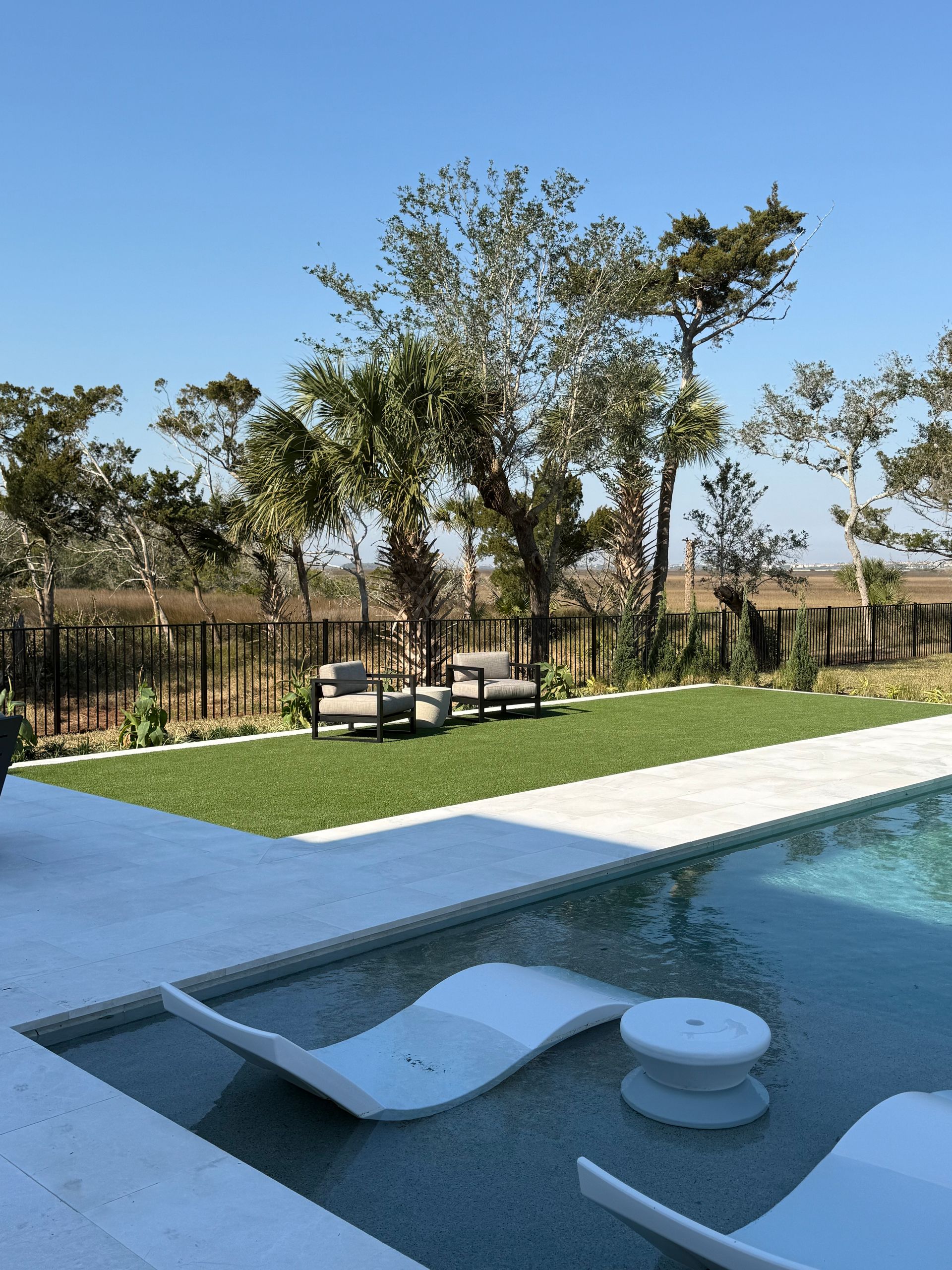 Poolside scene: Two chairs on a green lawn, trees in the background. Sunlight reflects on the water.