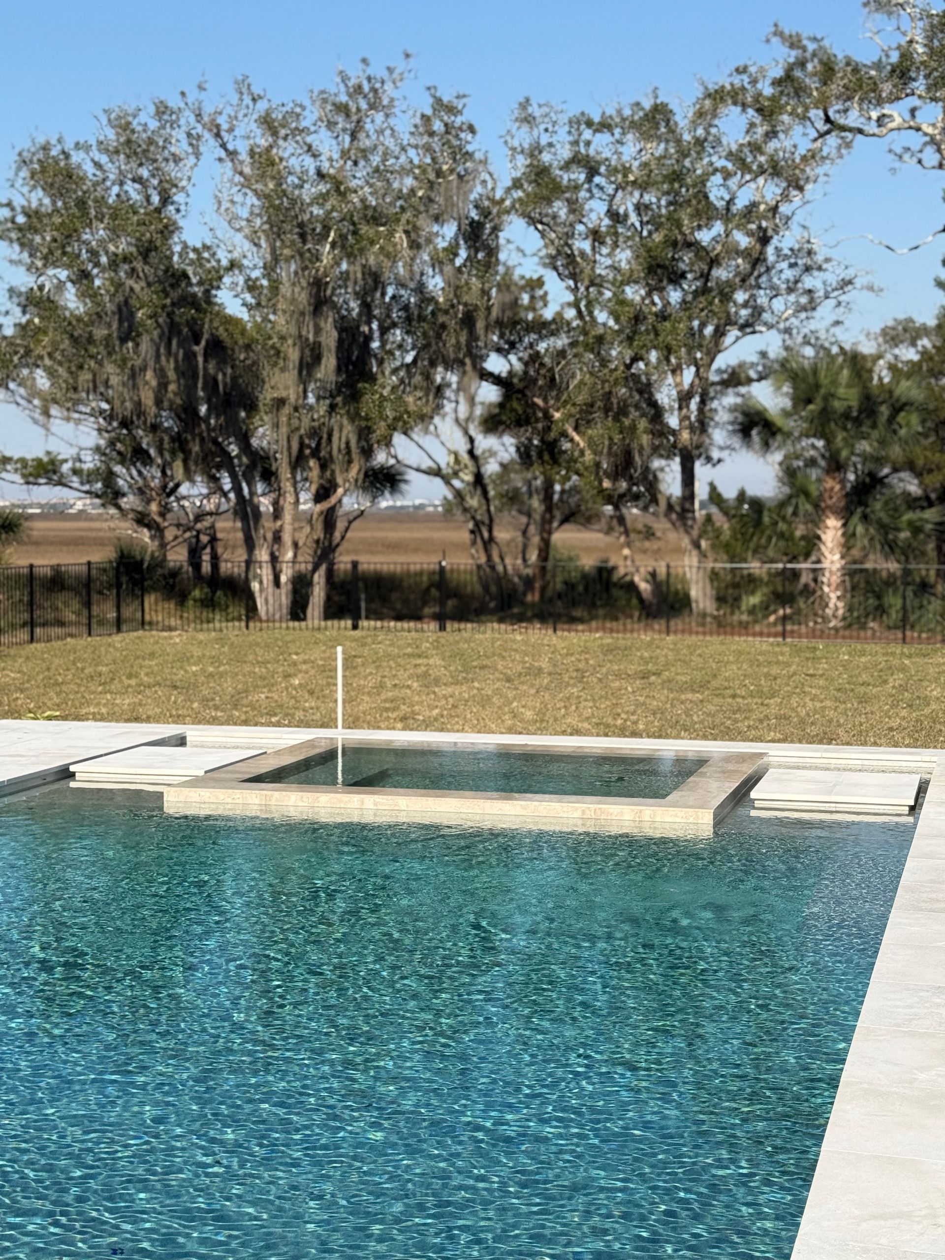 Pool with blue water and a square spa, grassy lawn, and trees under a blue sky.