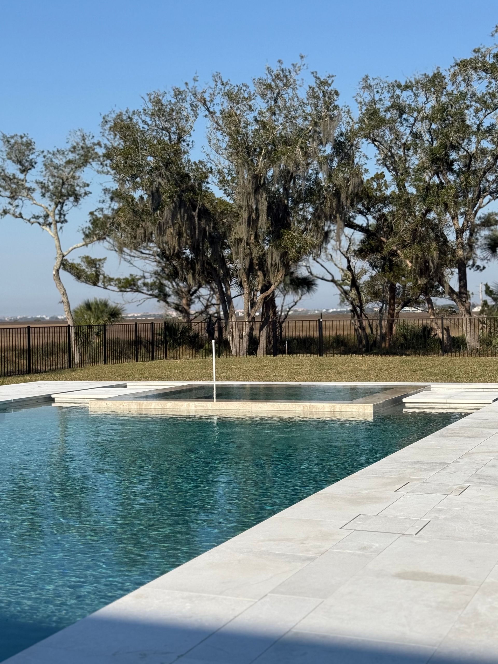 Swimming pool with blue water, white stone border, and trees in the background under a clear, blue sky.