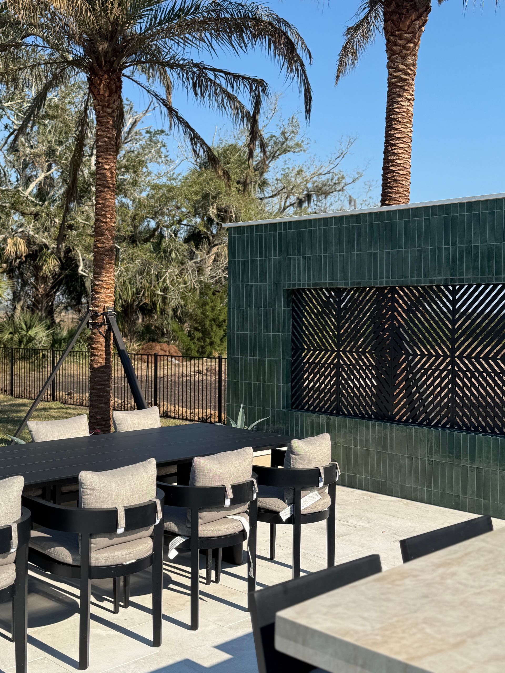 Outdoor dining area with a black table and chairs against a green tiled wall, palm trees in the background.