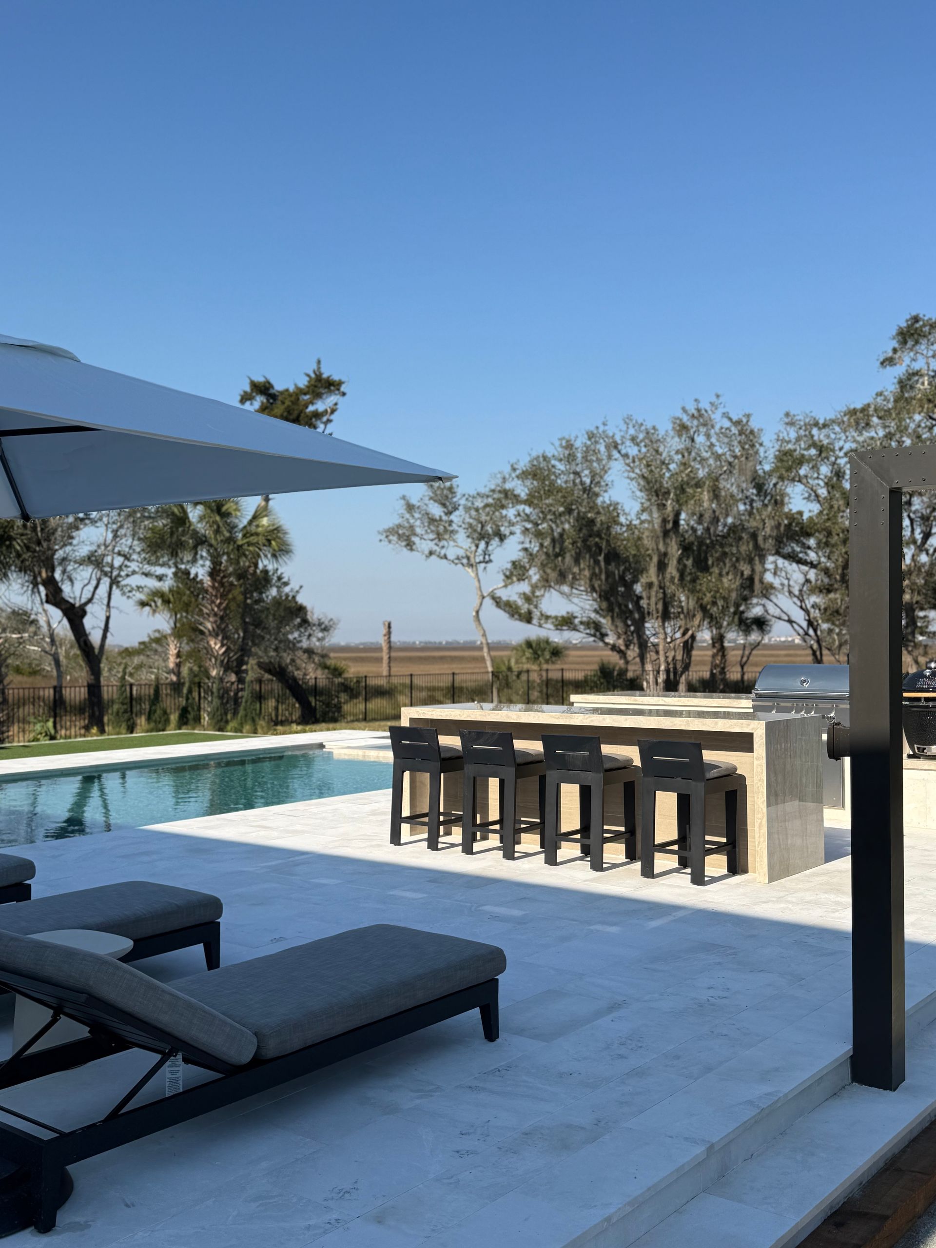 Poolside outdoor kitchen with chairs, umbrella, and a clear blue sky.