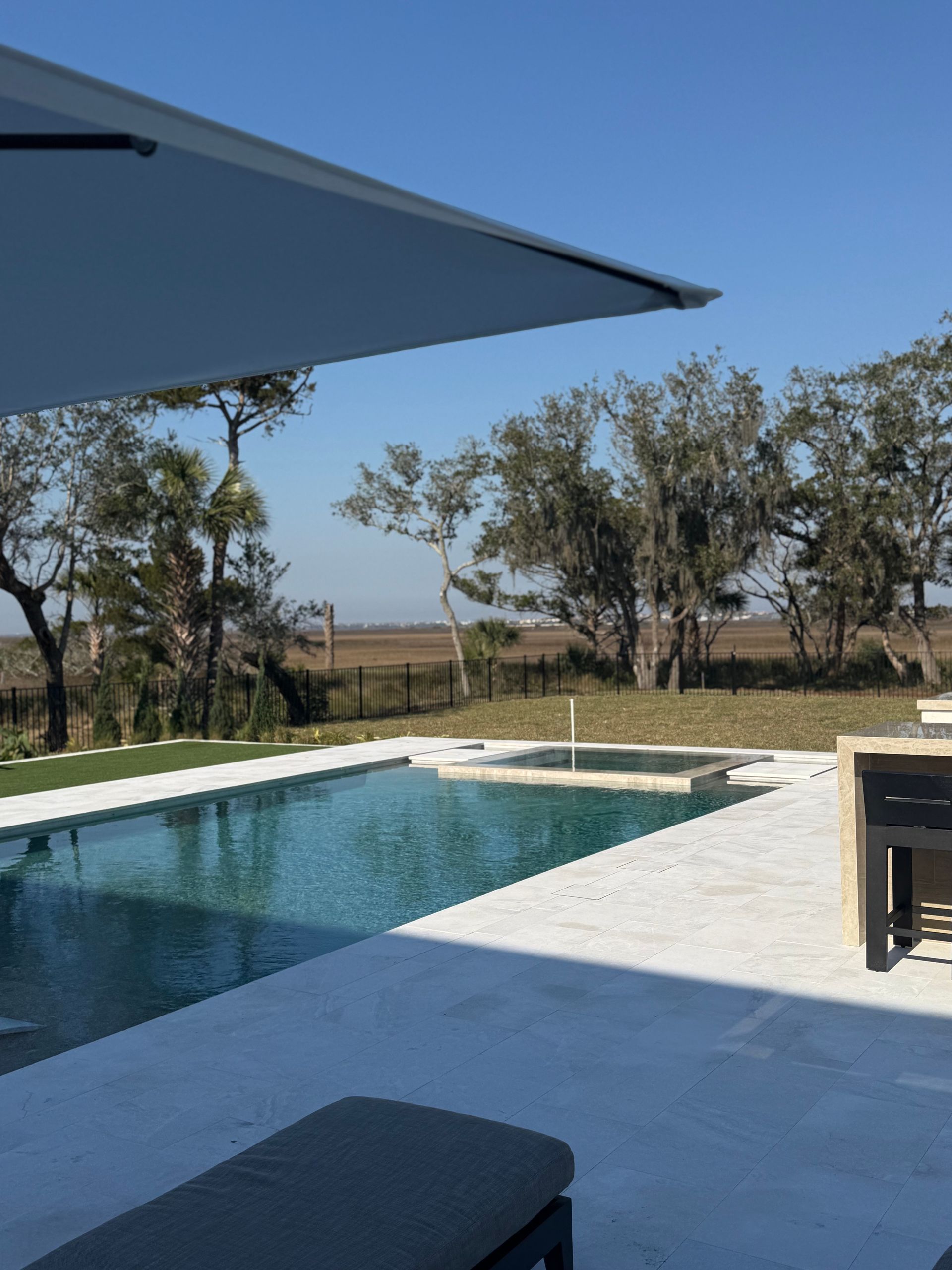 Pool with umbrella, jacuzzi, and field in background under a clear blue sky.