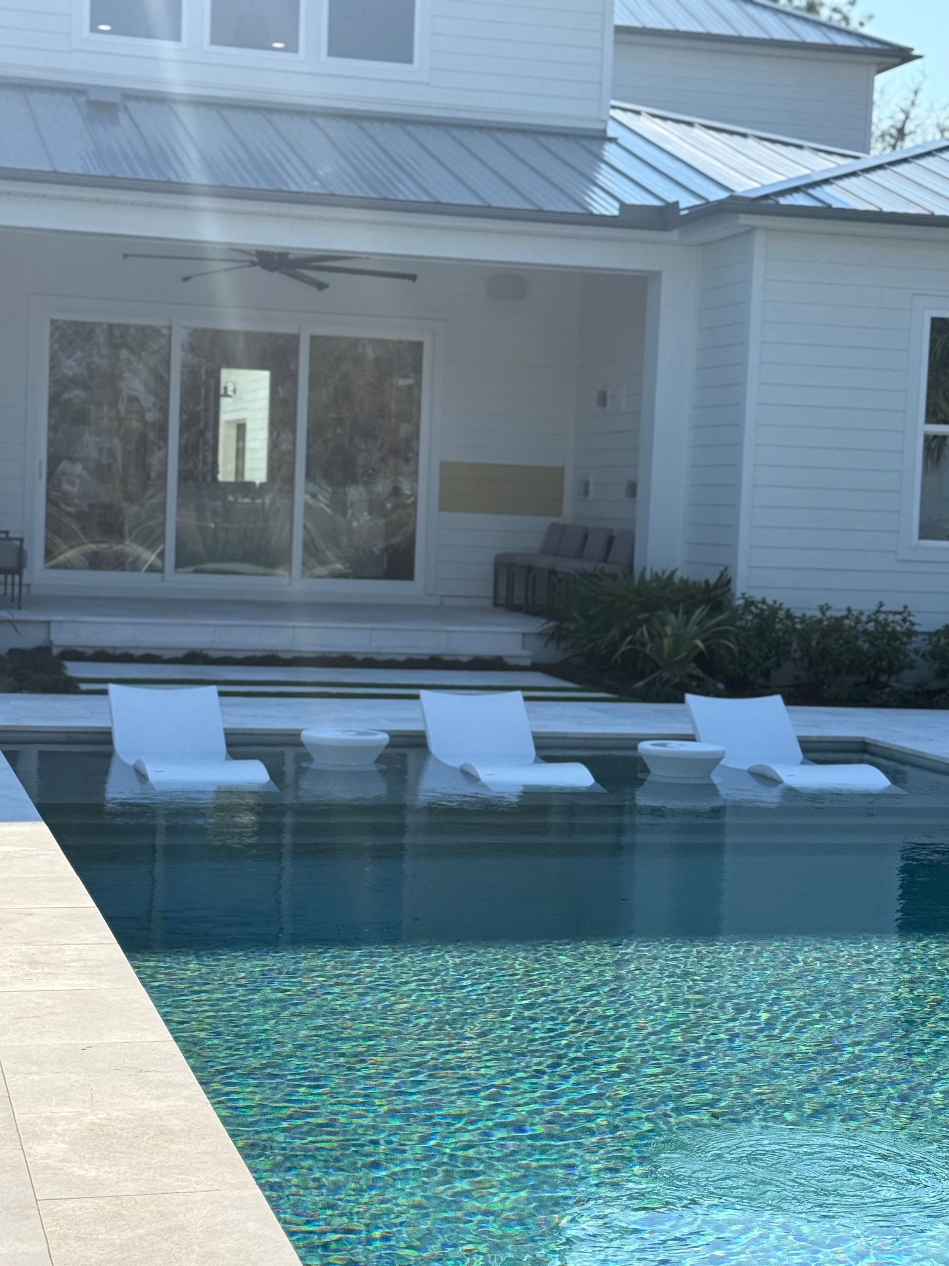 Pool with white lounge chairs, beside a white house with a patio and glass doors.