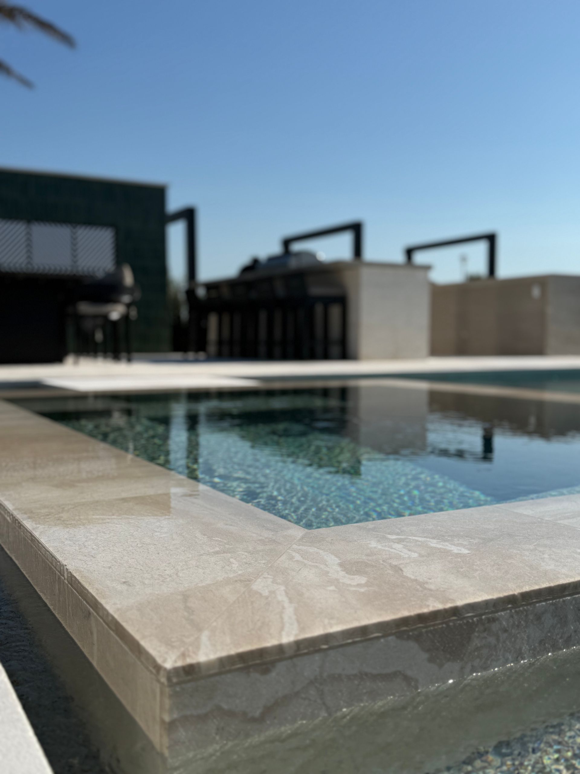 Pool with stone coping, overlooking outdoor kitchen and clear blue sky.