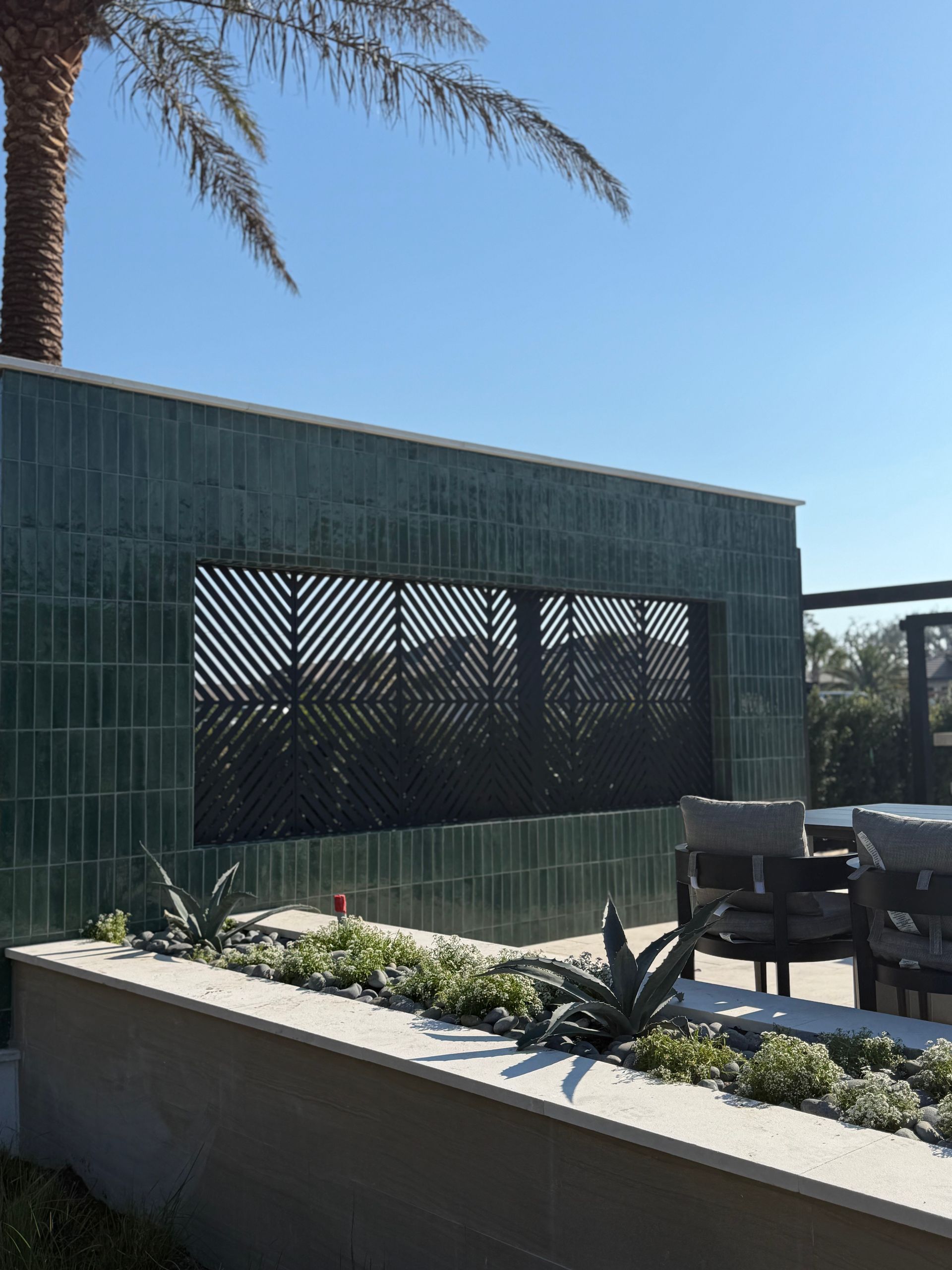 Green tiled wall with a lattice window, plants, and outdoor seating on a sunny day.