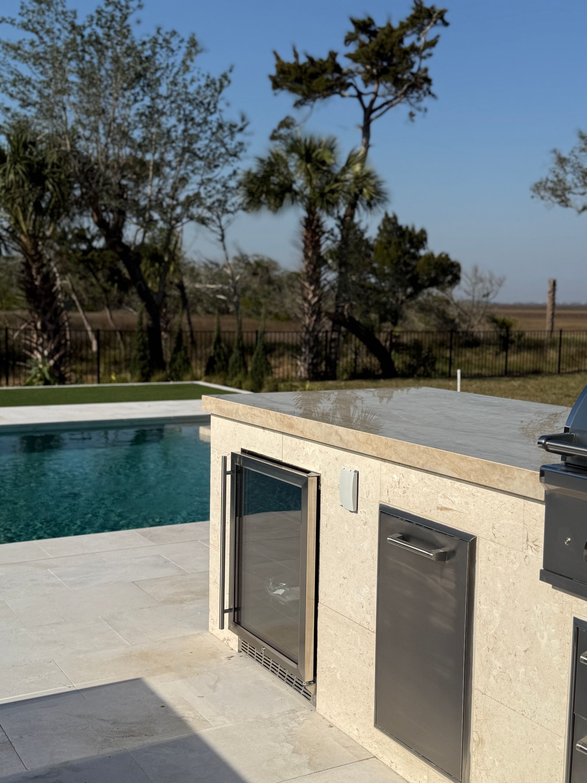 Outdoor kitchen next to a pool, with stainless steel cabinets and grill. Sunny, with trees and sky in background.