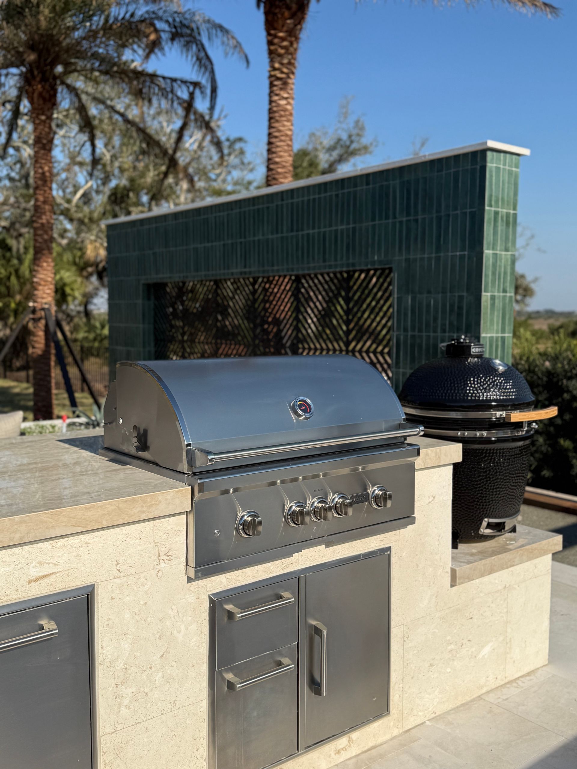 Outdoor kitchen with stainless steel grill, built-in storage, and ceramic egg smoker.  Green tiled wall in a sunny setting.