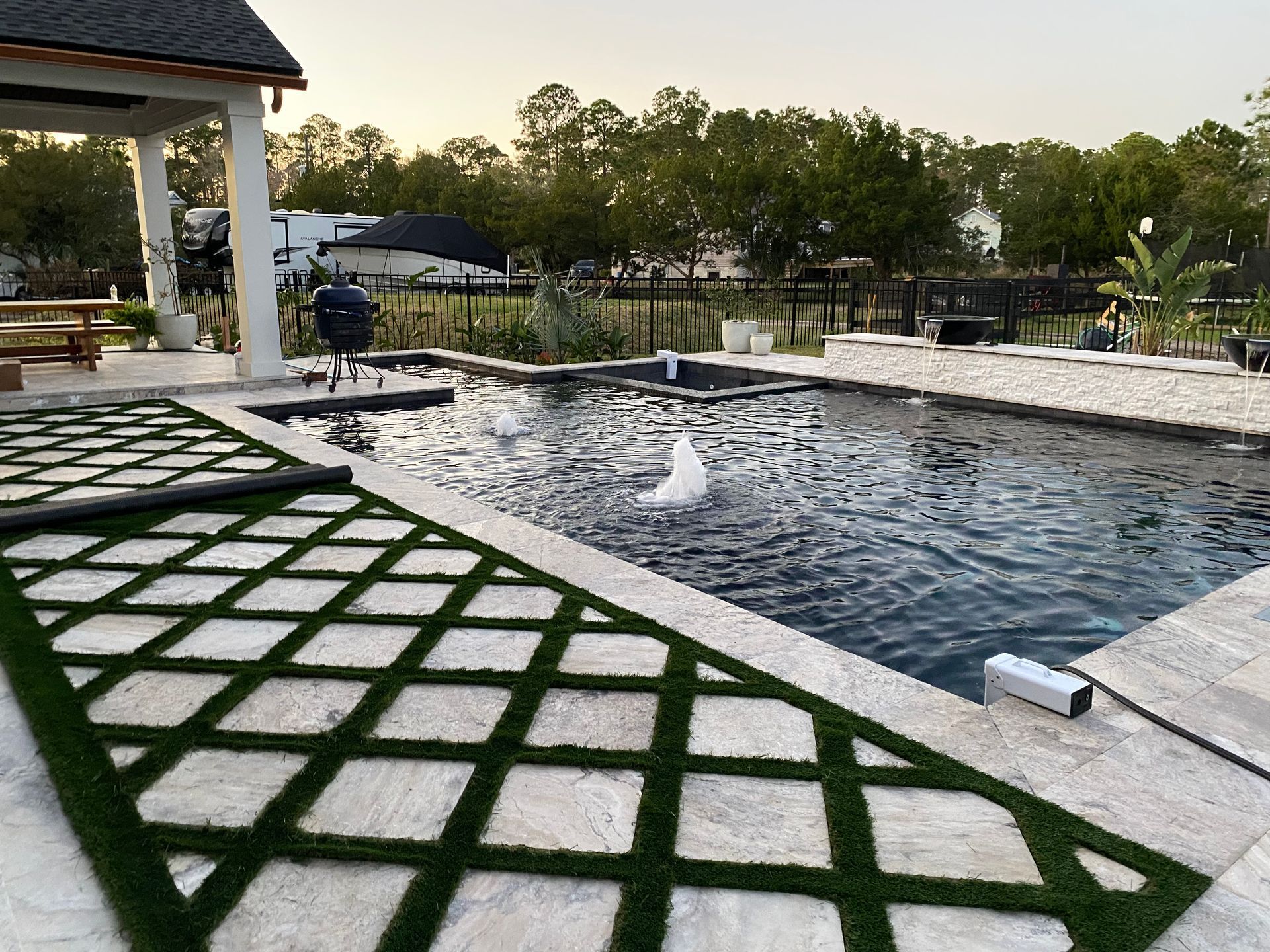 Patio with a pool, gazebo, and diamond-patterned lawn. A person grills food.