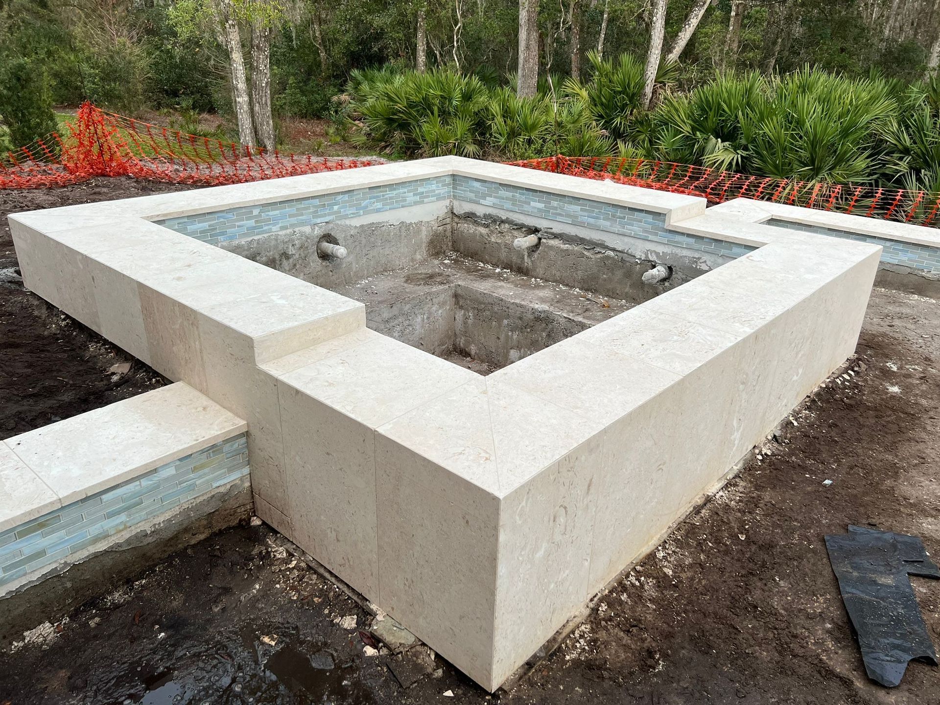 Empty, square-shaped swimming pool under construction with light beige block walls, and surrounding landscaping.