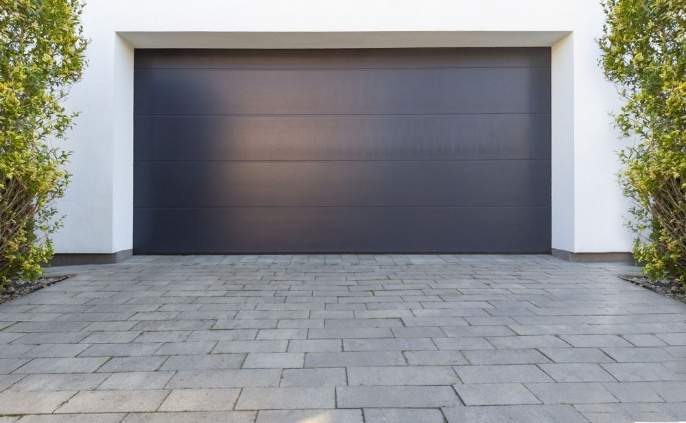 Dark gray garage door on a white building, with brick driveway and bushes.