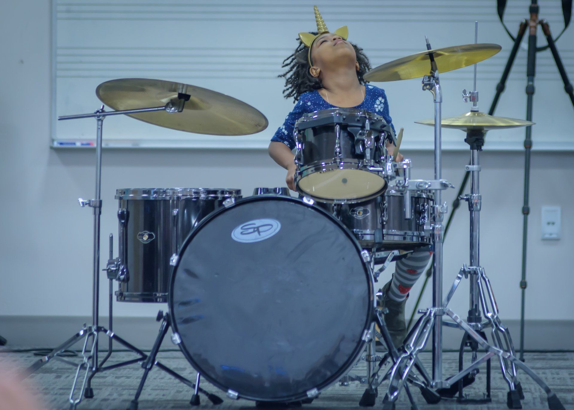 A young girl wearing a unicorn horn is playing drums.