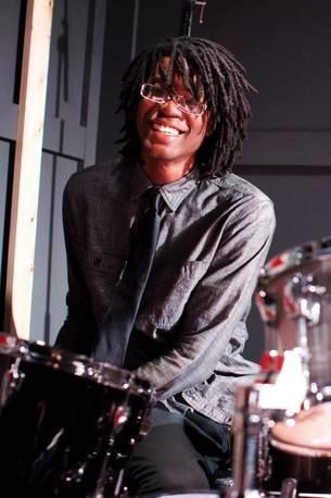 A man with dreadlocks is playing drums and smiling.