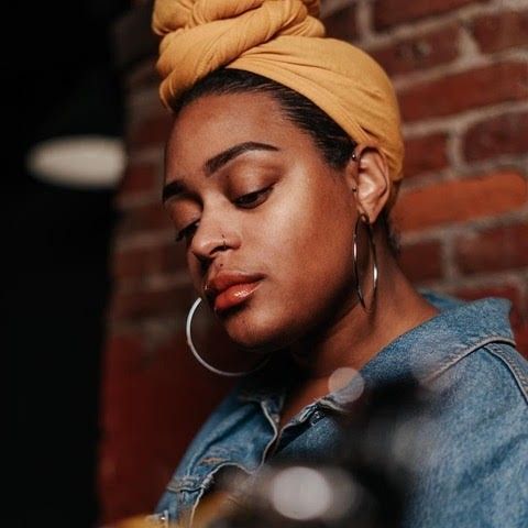 A woman wearing a yellow turban and hoop earrings is sitting in front of a brick wall.