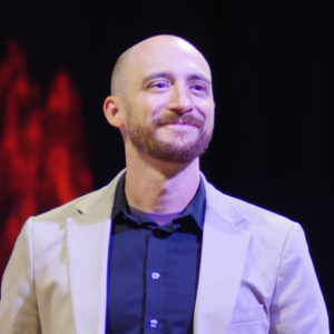 A bald man with a beard wearing a tan jacket and a black shirt