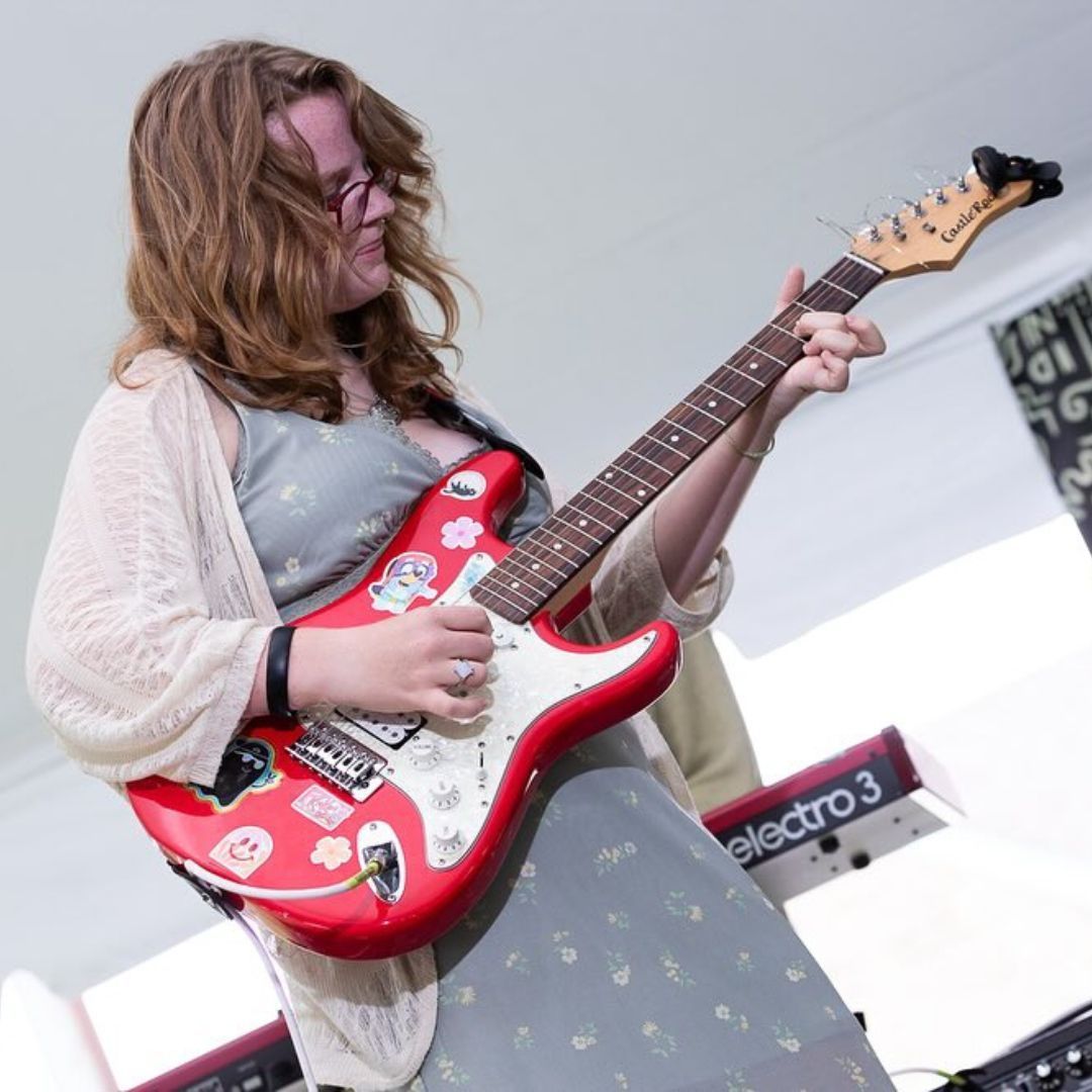 A woman is playing a red electric guitar in front of an electro 3 keyboard