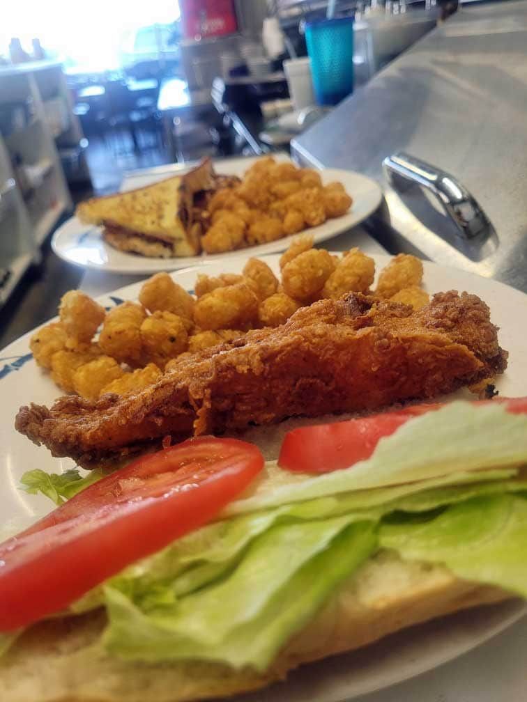 Fried fish sandwich with lettuce, tomato, and tater tots on a white plate; other plate has a grilled sandwich and tots.