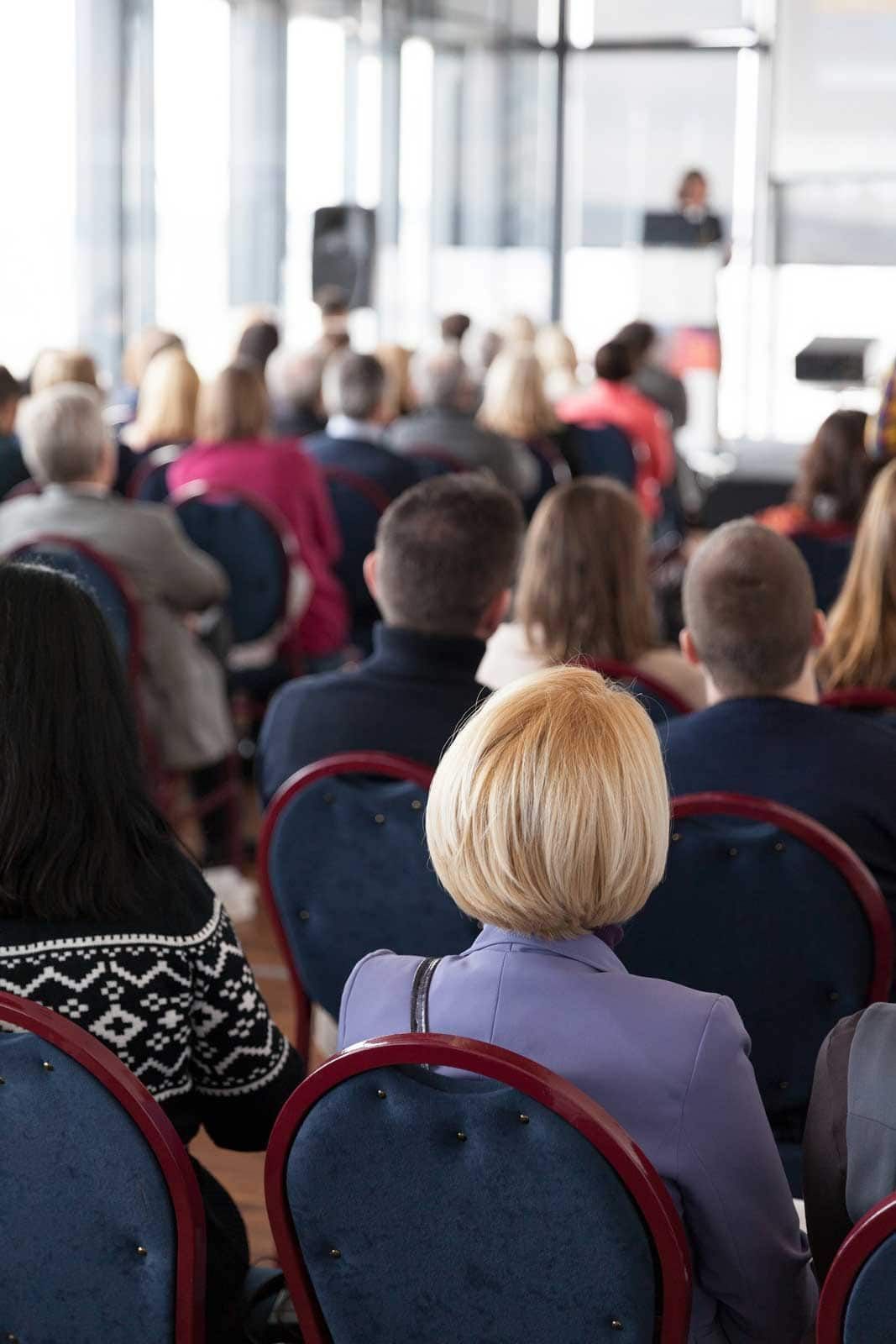 Audience seated in chairs, facing a speaker at a podium. Indoor setting, natural light.