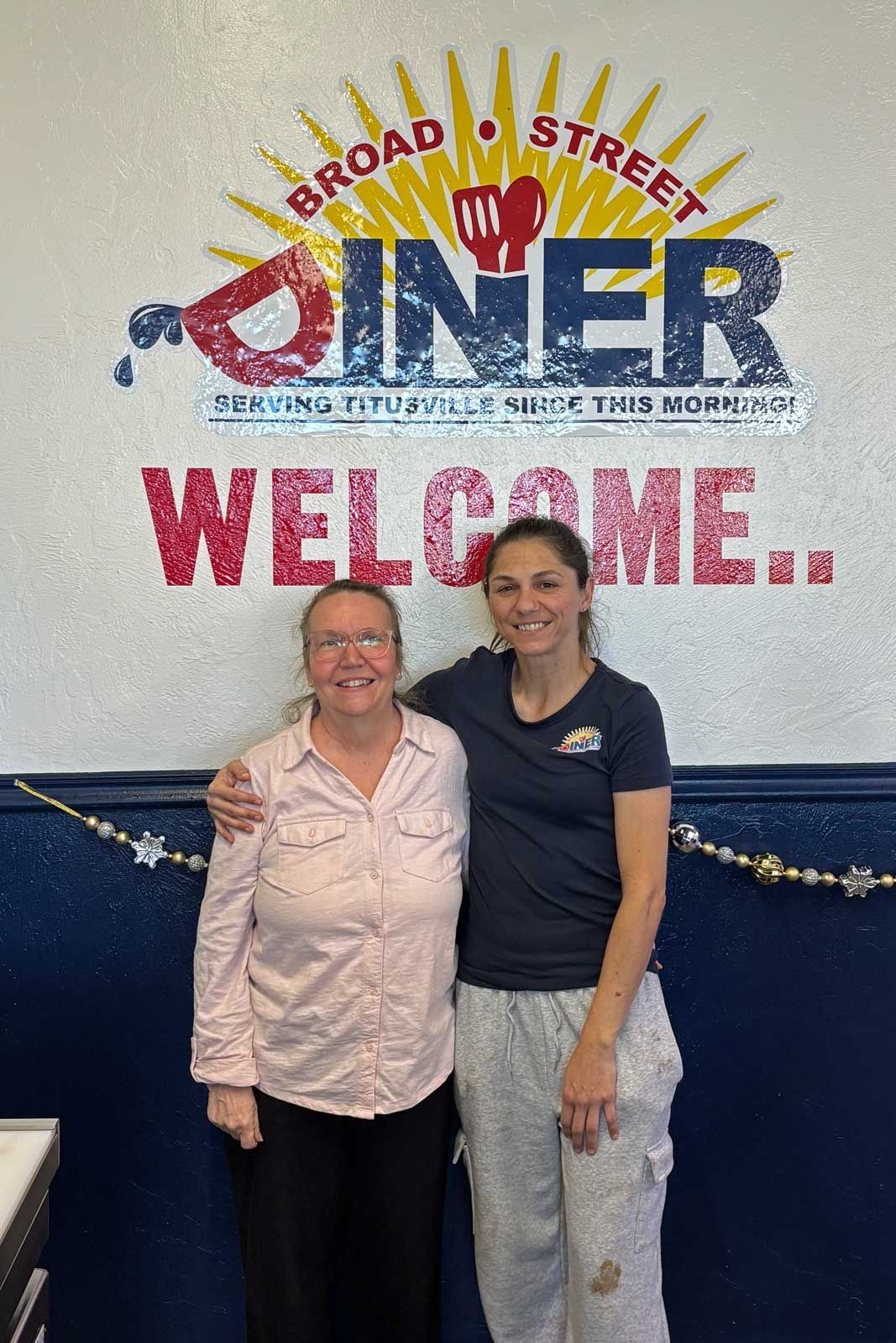 Two women pose in front of a Broad Street Diner sign. One has arm around the other.
