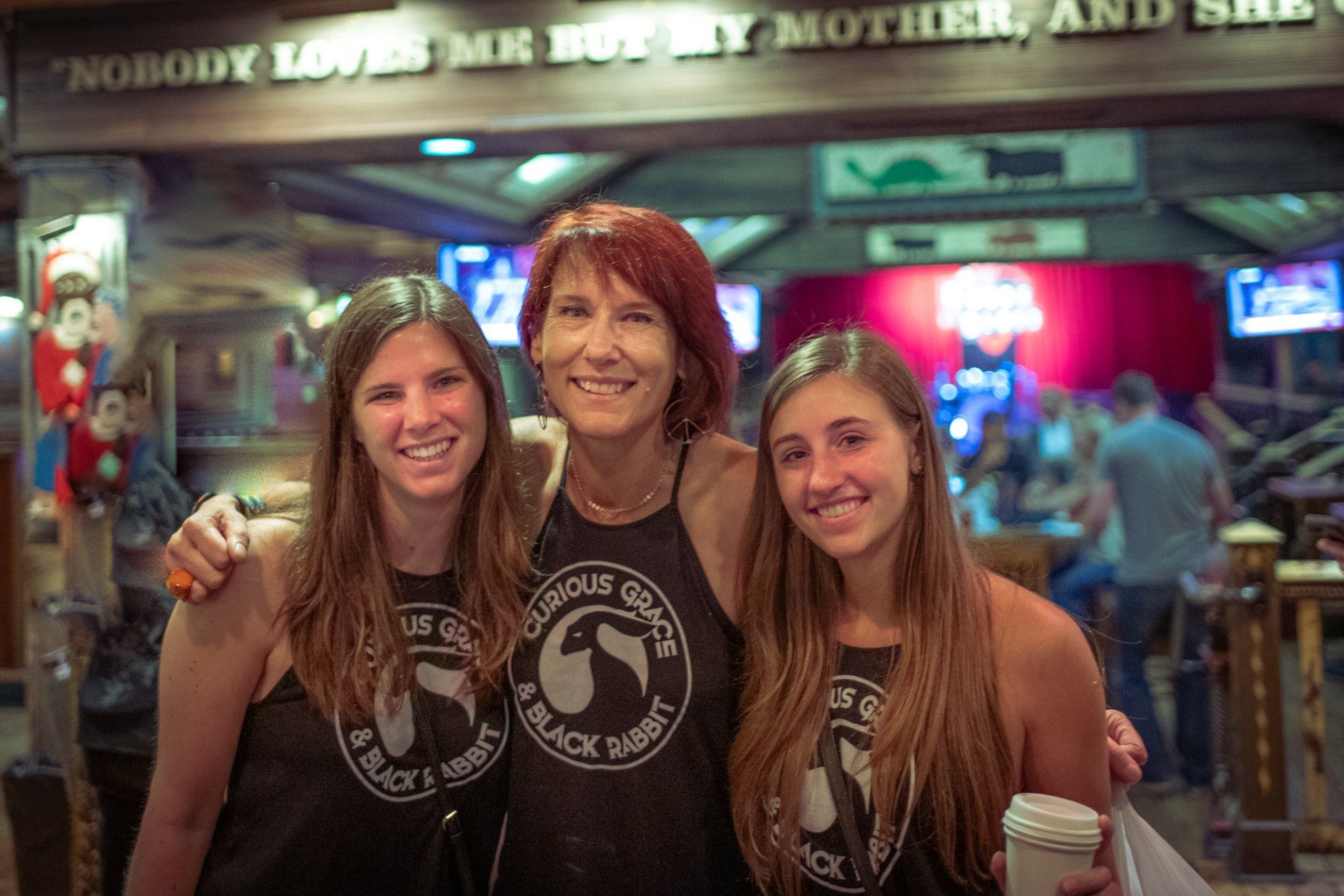 Three women are posing for a picture in front of a sign that says `` nobody loves me but my mother and she ''.