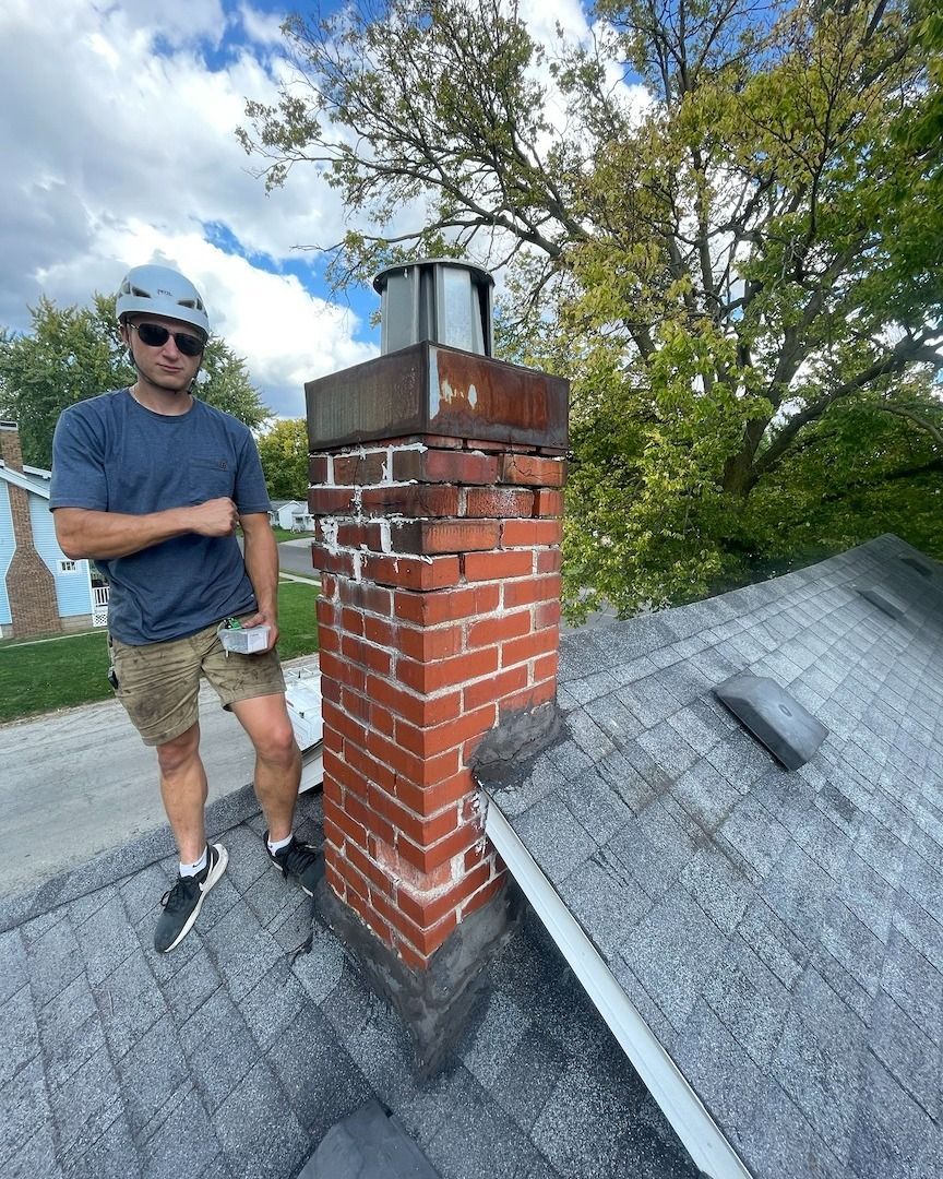 A man is standing on top of a roof next to a brick chimney.