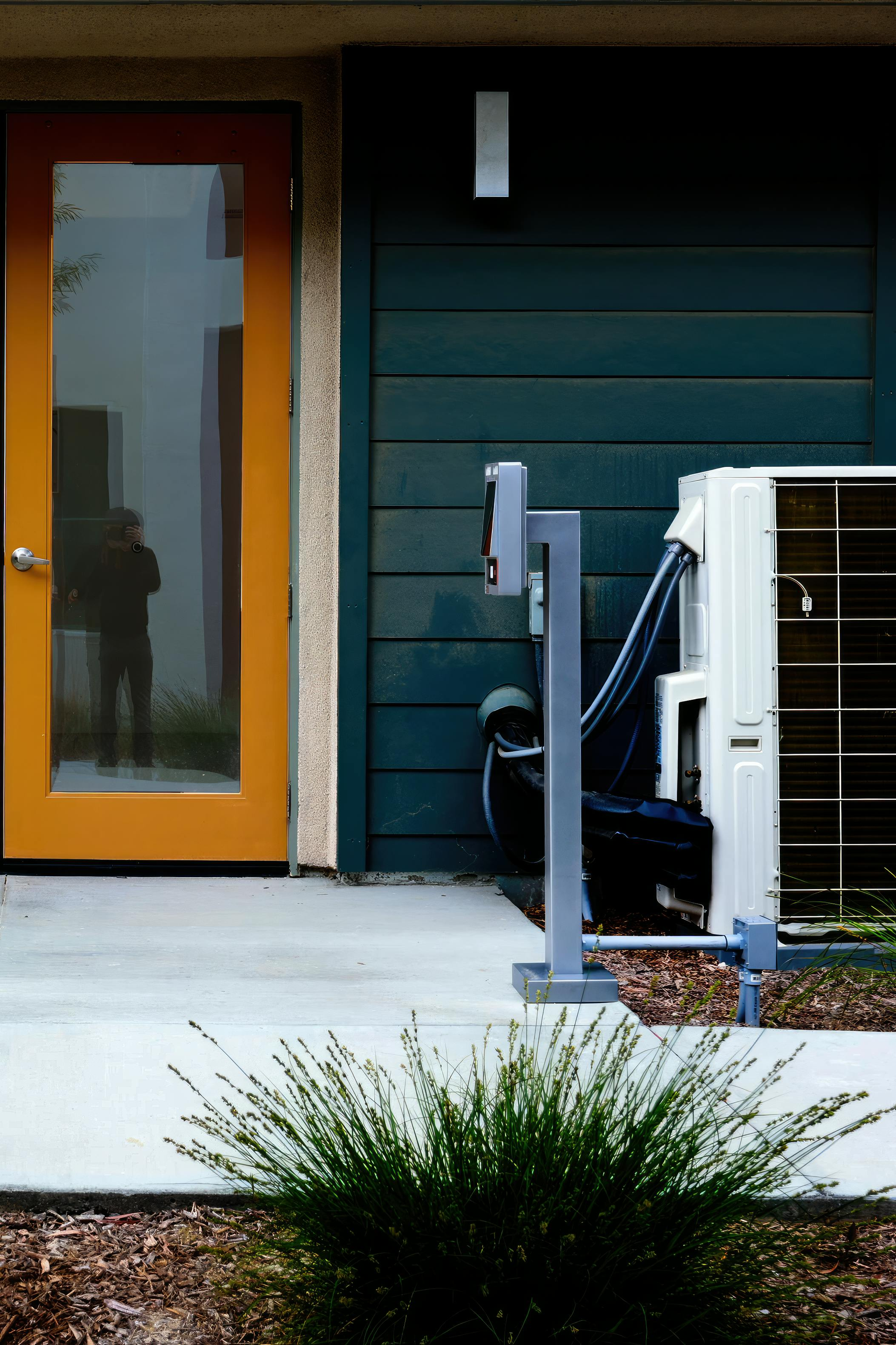 Front porch with orange door, dark siding, and a wheelchair ramp railing beside a small bush.