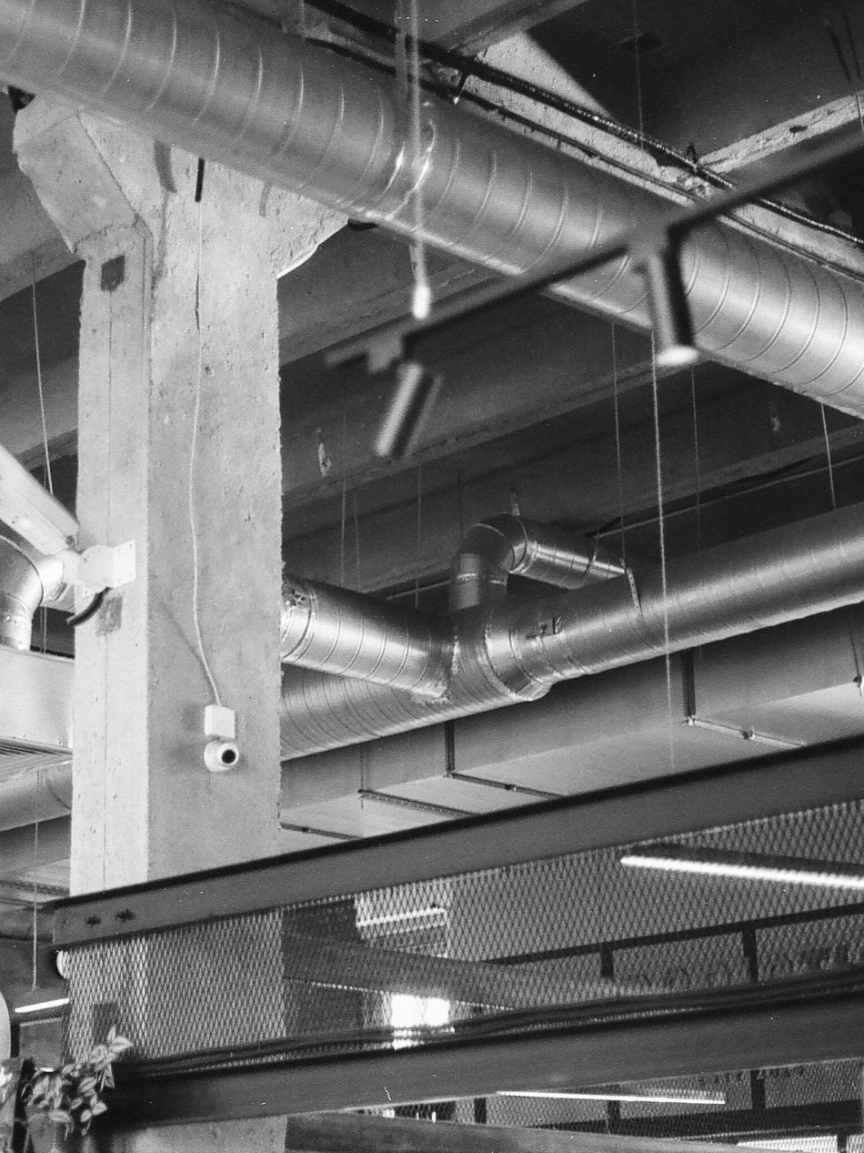Industrial ceiling with exposed ducts, pipes, and a concrete support column above a mesh barrier.