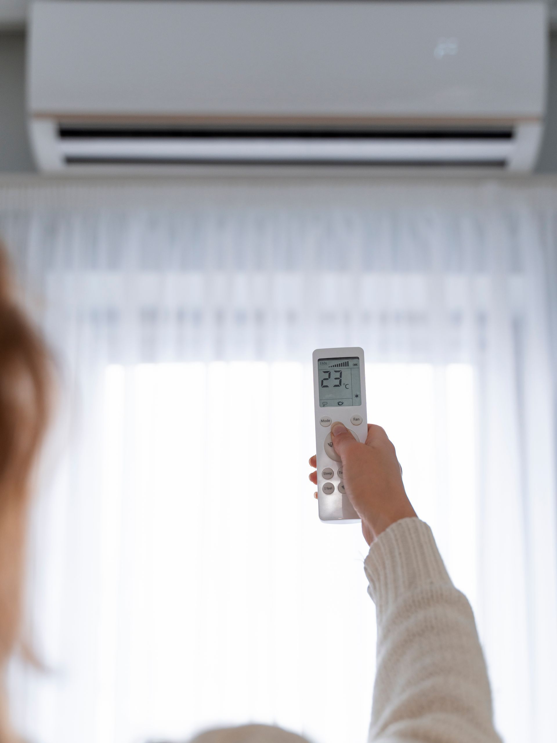 Hand holding a remote toward a wall-mounted air conditioner above a bright window with curtains