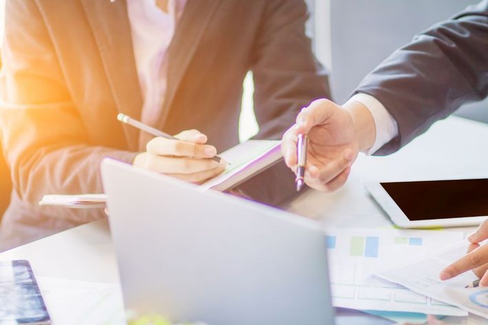 Business people in suits reviewing documents and using a laptop and tablet at a table, bright light.