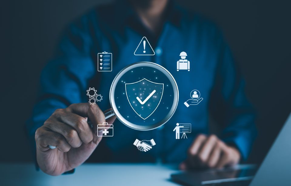 Person holds magnifying glass over a shield symbol surrounded by security icons.