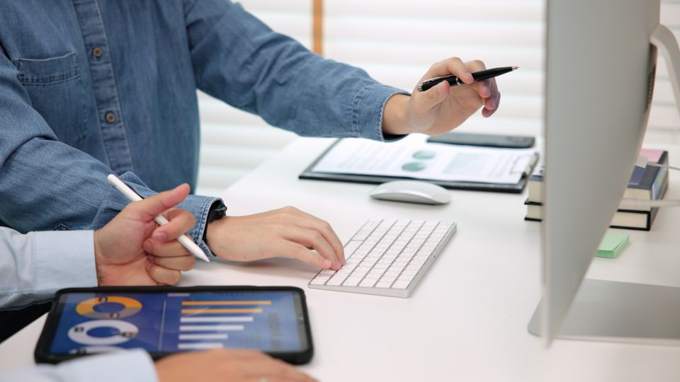 Two people collaborating at a desk, one pointing at a screen and the other using a tablet with charts.
