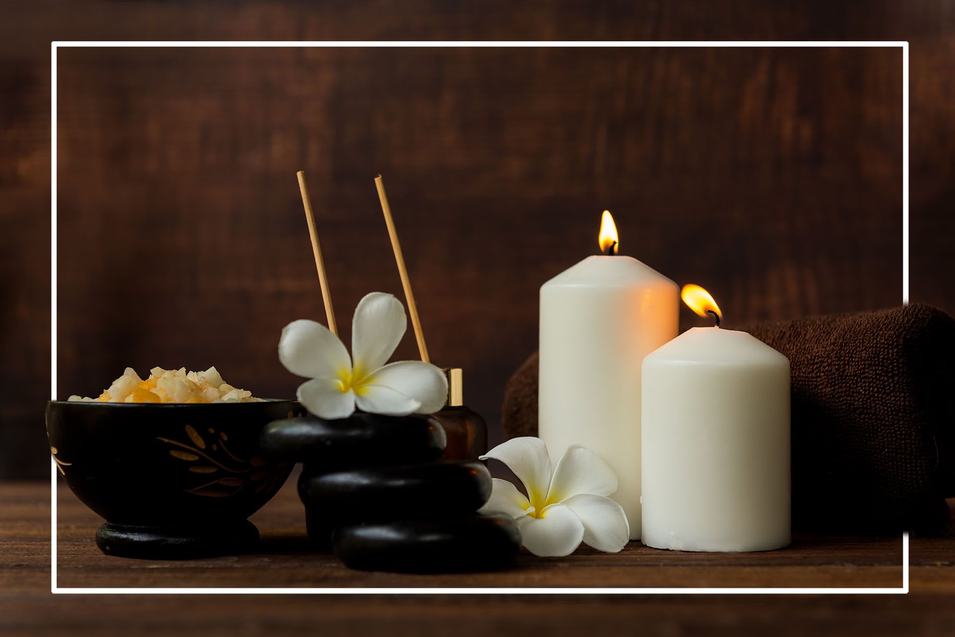 Spa setting: candles, black stones, flowers, incense, and bowl of salt against a wood backdrop.