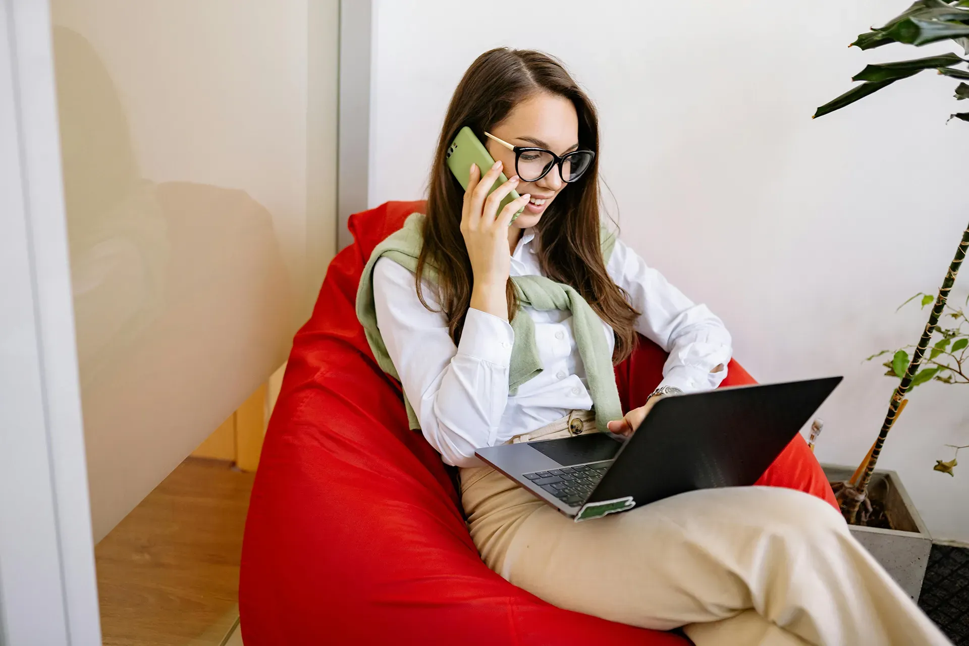 Woman on phone, using laptop, sitting in red beanbag chair.