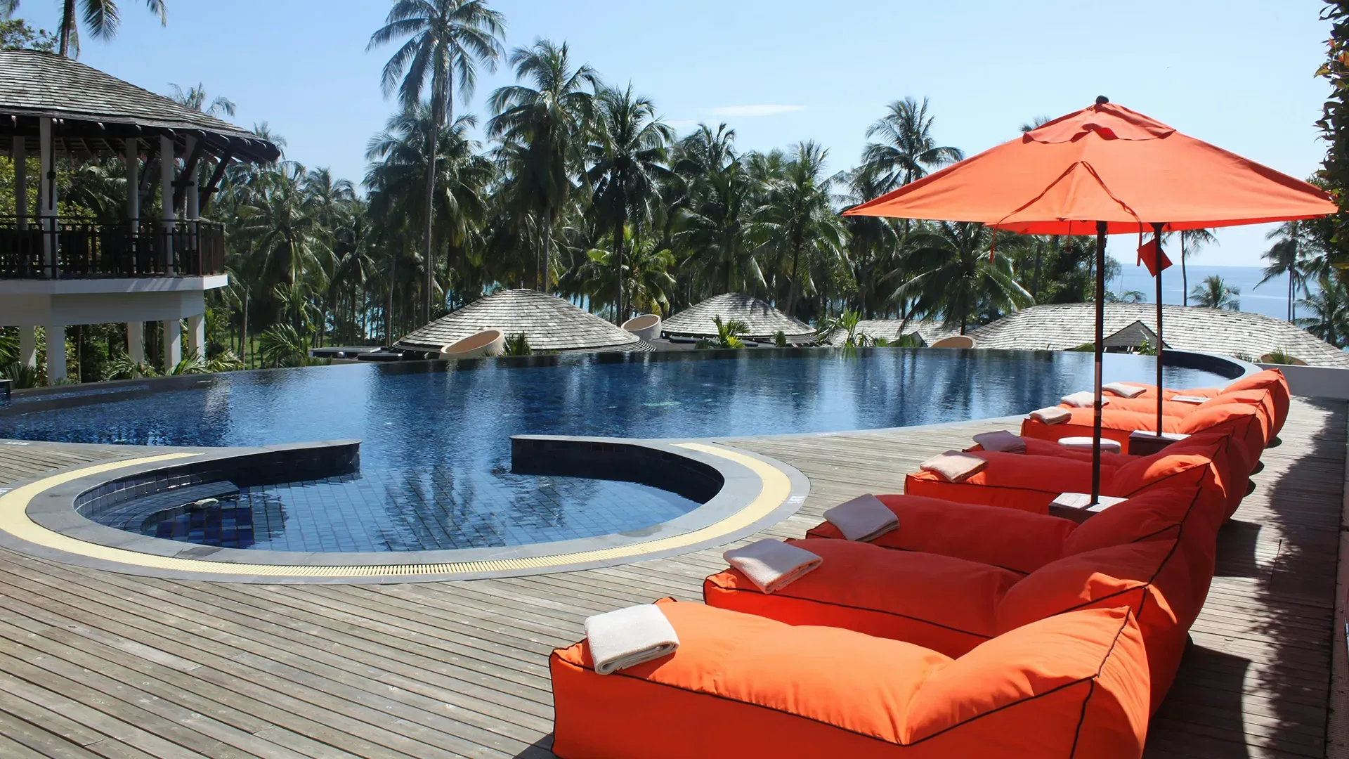 Infinity pool with orange lounge chairs, orange umbrella, and palm trees under a sunny sky.