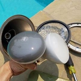 A pool repair person is holding a pool light bulb in their hand next to a swimming pool.